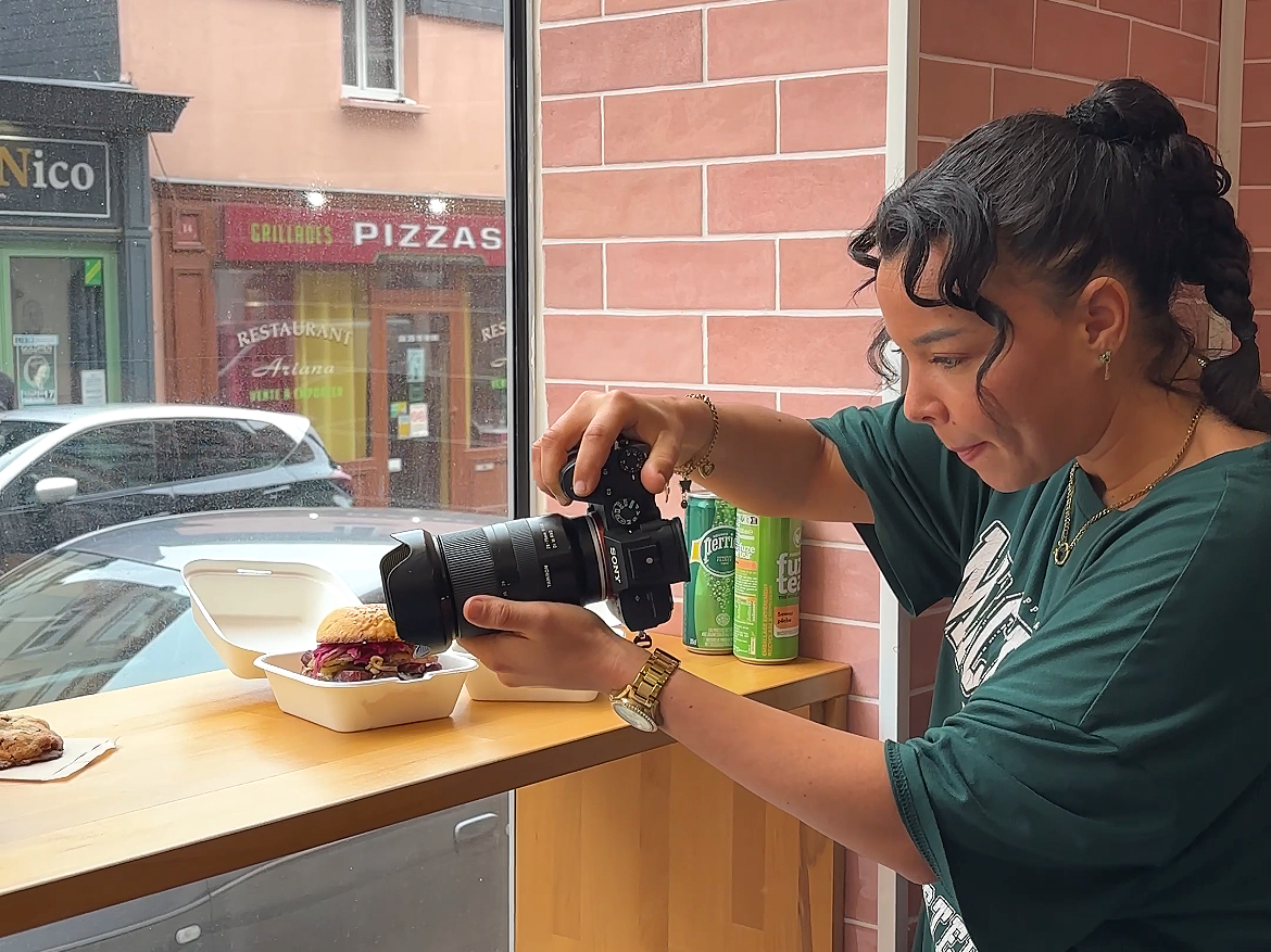 Une femme prend une photo d'un sandwich dans un restaurant. Il y a des canettes de boissons derrière elle et une voiture visible à l'extérieur par la fenêtre.