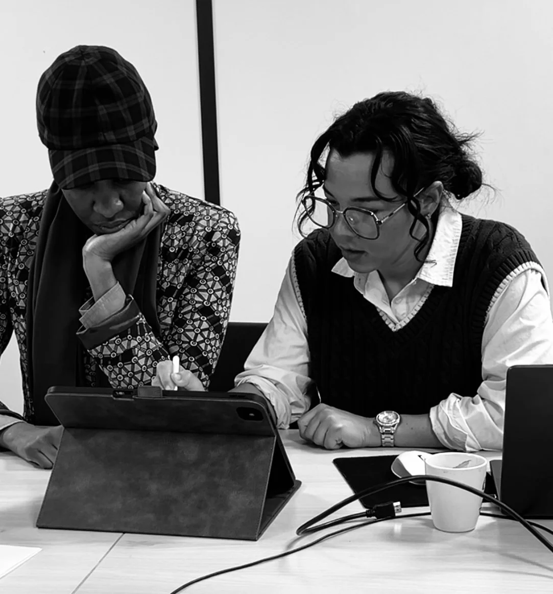 Deux personnes travaillent ensemble à une table, l'une tient une tablette, l'autre regarde un appareil électronique. En face, une tasse blanche et un ordinateur portable sont visibles.