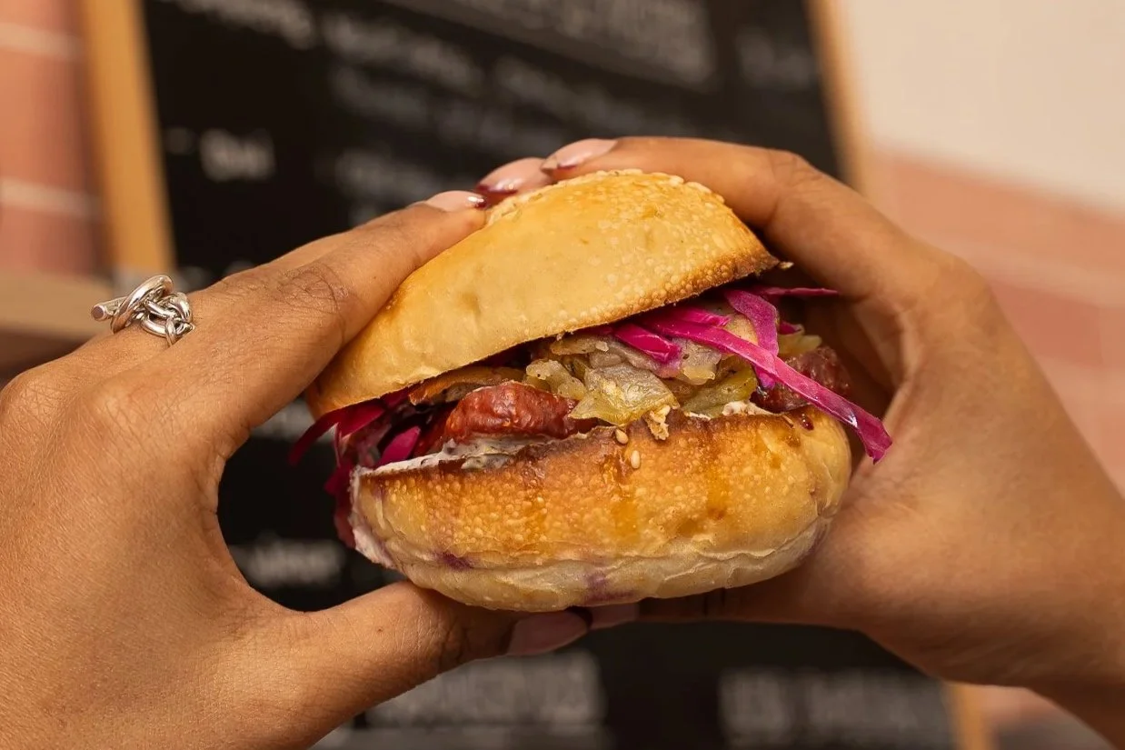 Une personne tenant un sandwich avec du pain, de la viande, de la choucroute et de la salade, devant un tableau noir avec des écrits