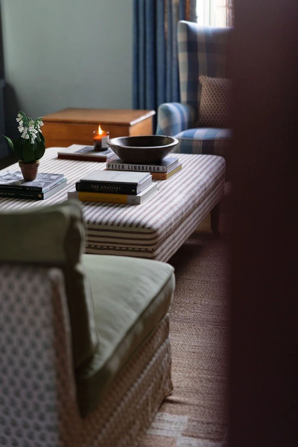 A cozy living room corner with a striped fabric-covered ottoman holding stacked books, a black bowl, and a lit candle. A potted plant with white flowers decorates the ottoman. An armchair with patterned fabric is partially visible in the foreground, 