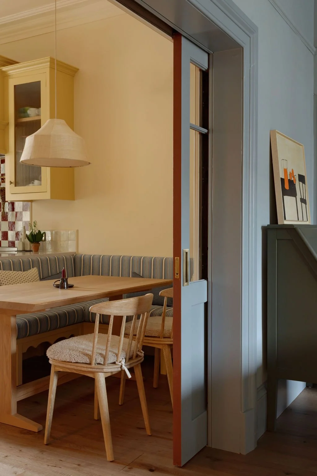 View into a cozy dining area with wooden furniture, striped bench seating, a wall with a small kitchen area, and artwork on the wall