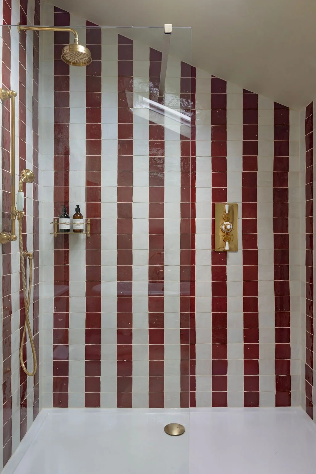Modern shower with red and white striped tiles, a gold showerhead, a handheld shower, built-in soap shelves, and a gold control panel.