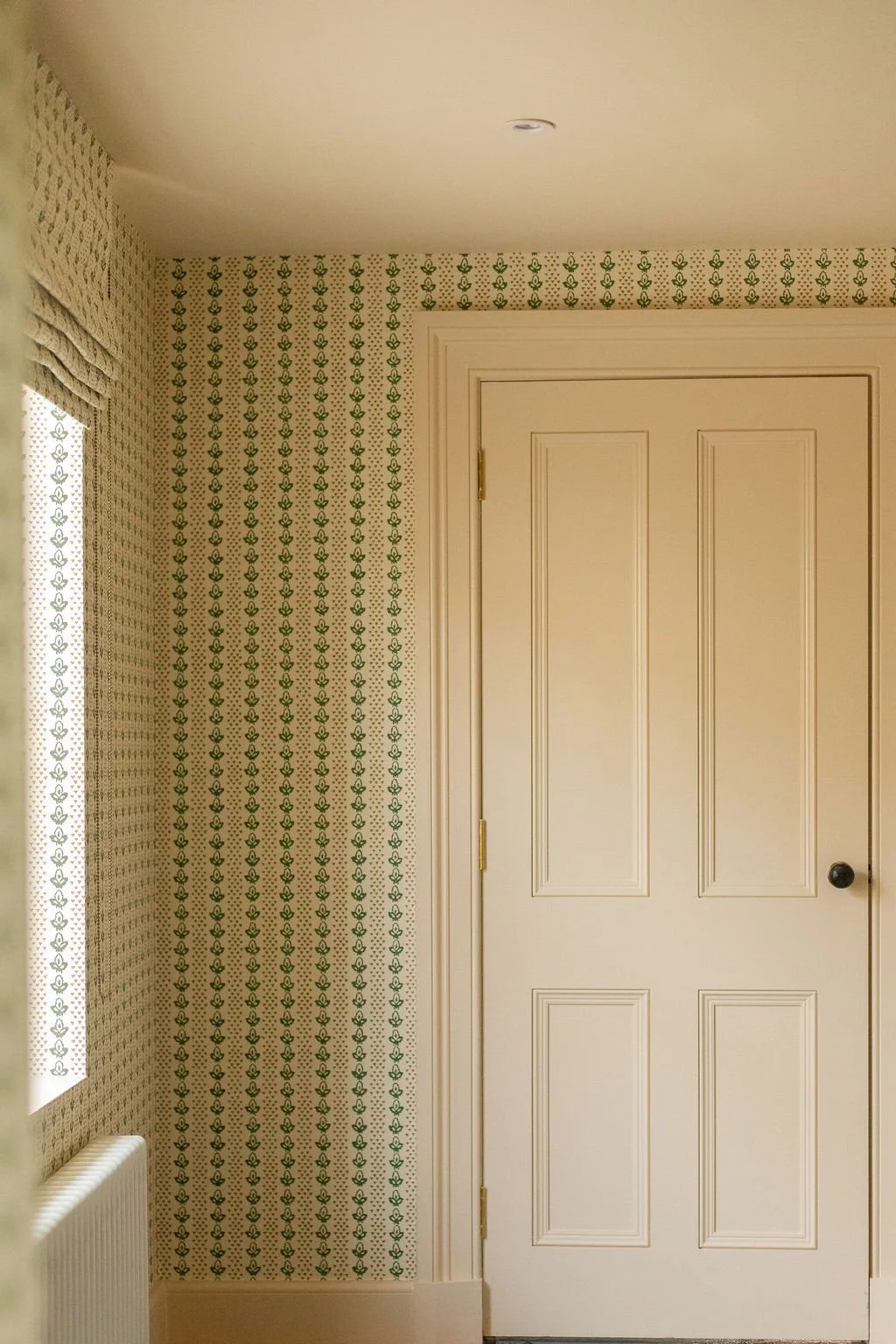 Interior view of a room corner with patterned wallpaper in green and beige, a closed white door, and part of a window with curtains.