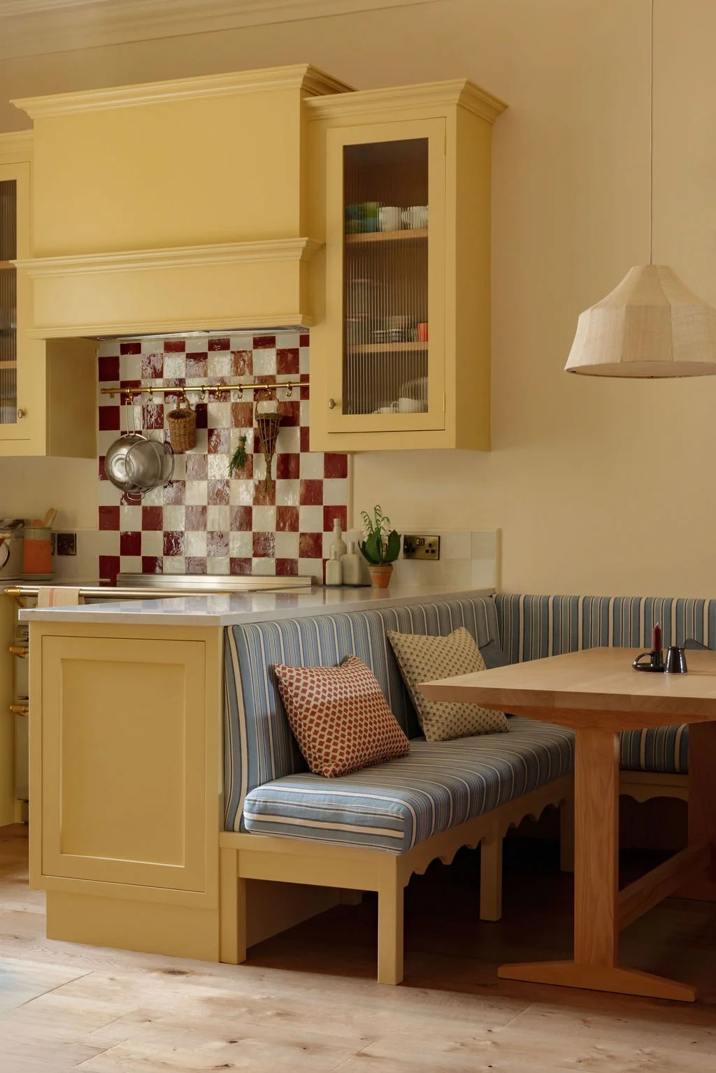 Cozy kitchen with yellow cabinetry, checkered red and white backsplash, striped seating with pillows, and a wooden table with a pepper shaker and salt shaker.