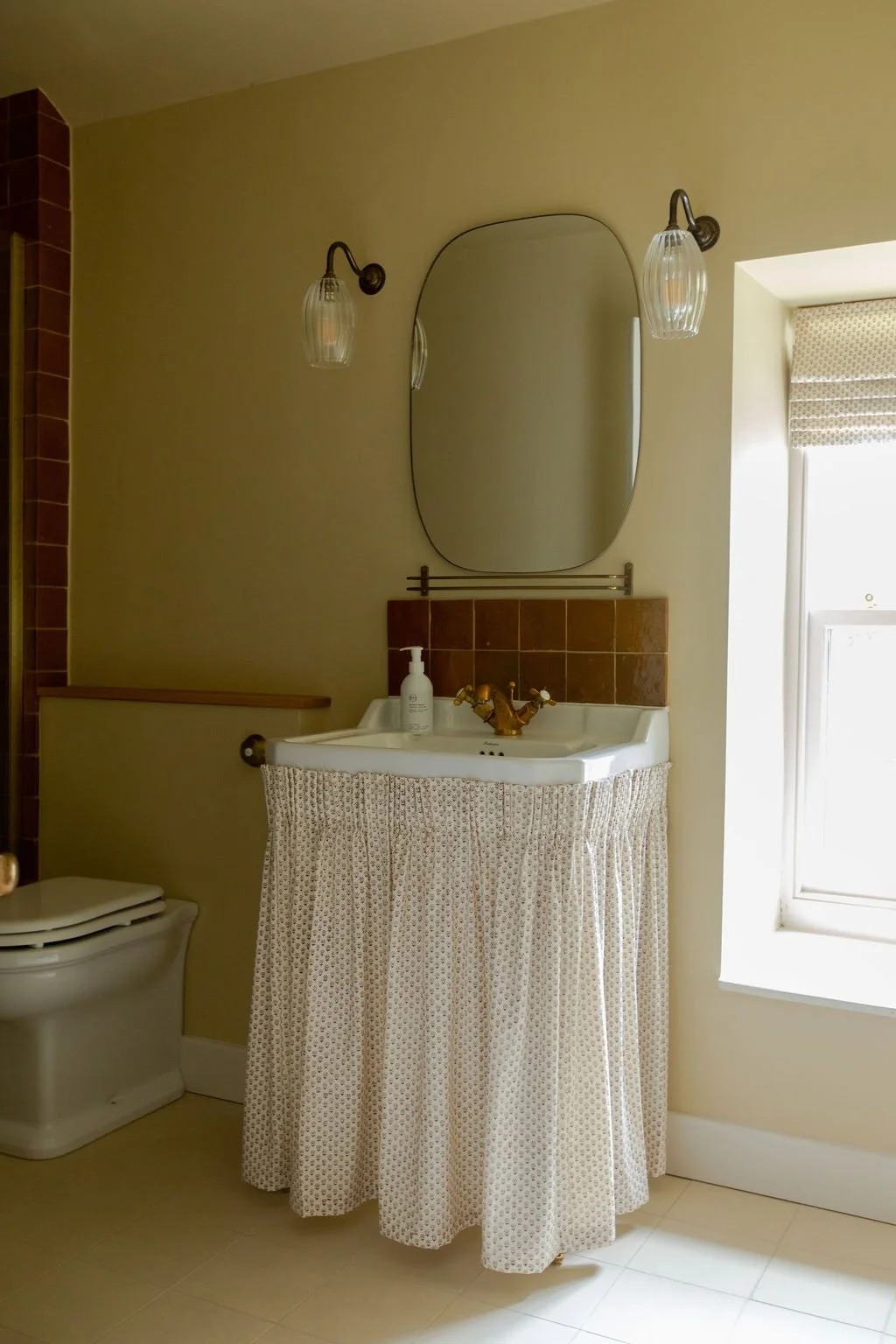 Bathroom with a small sink covered by a patterned curtain, a mirror above, a wall lamp, and a window with a shade.