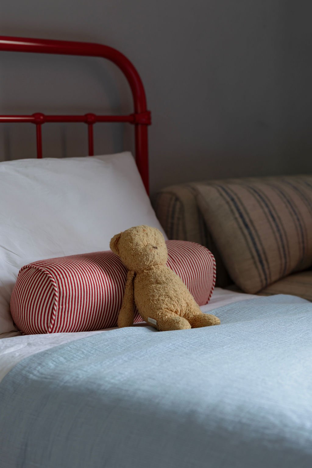 A cozy bed with white bedding, red striped bolster pillow, a worn stuffed teddy bear, and a sofa with striped upholstery in the background.