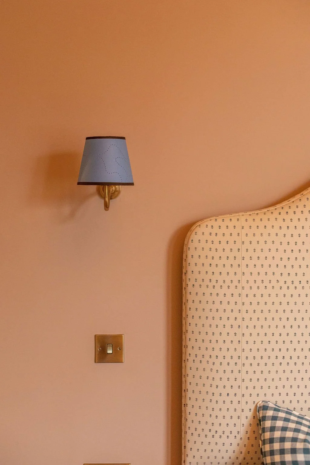 Close-up of a bedroom wall with a wall-mounted light fixture, part of a bed with a patterned headboard, and a pillow with a checkered pillowcase.