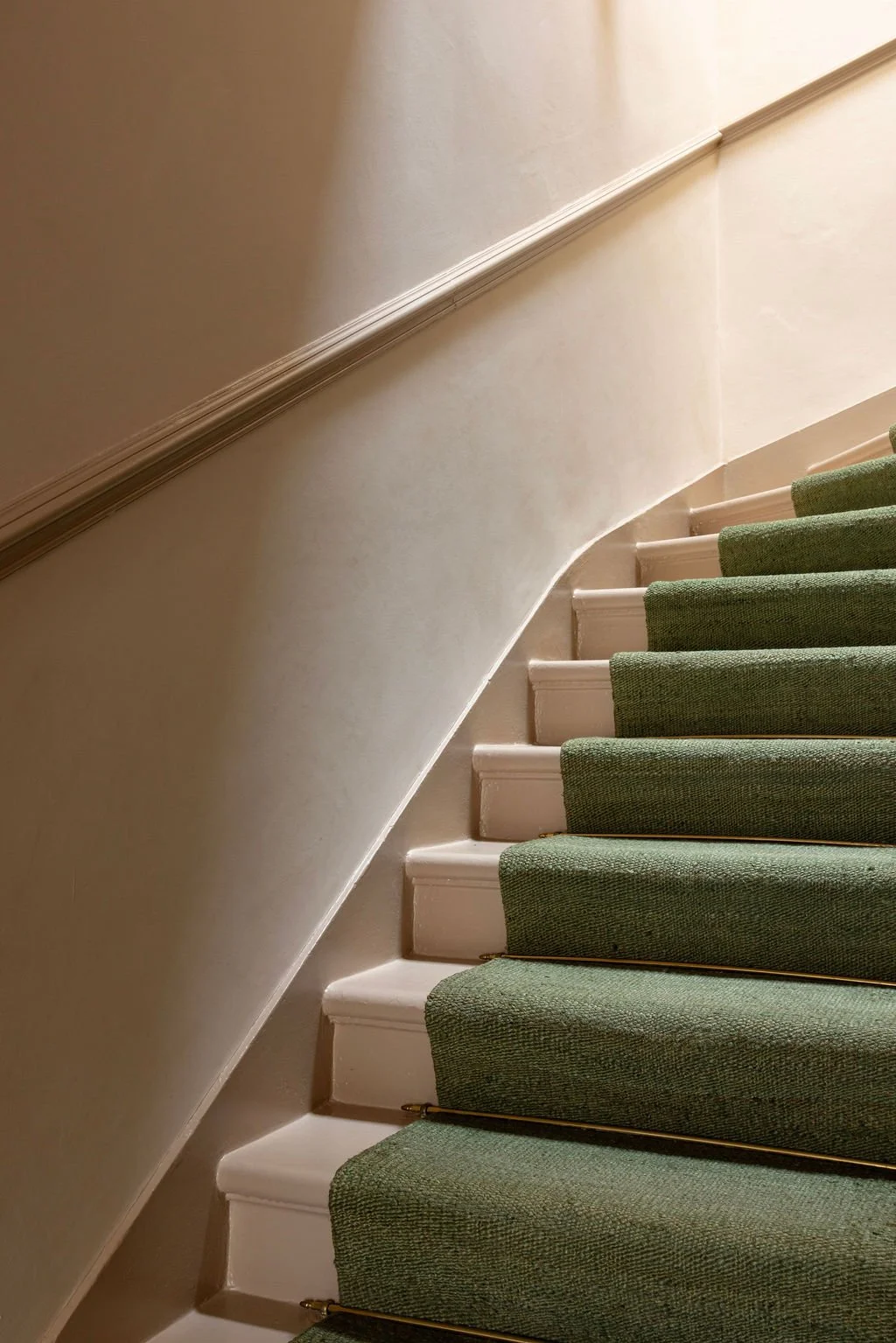Part of an indoor staircase with white-painted steps and green carpet runners, along with a beige wall and a decorative wooden handrail.