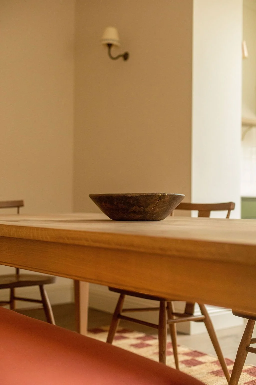 A brown ceramic bowl placed on a wooden dining table in a room with cream walls, wooden chairs, and a patterned rug on the floor.