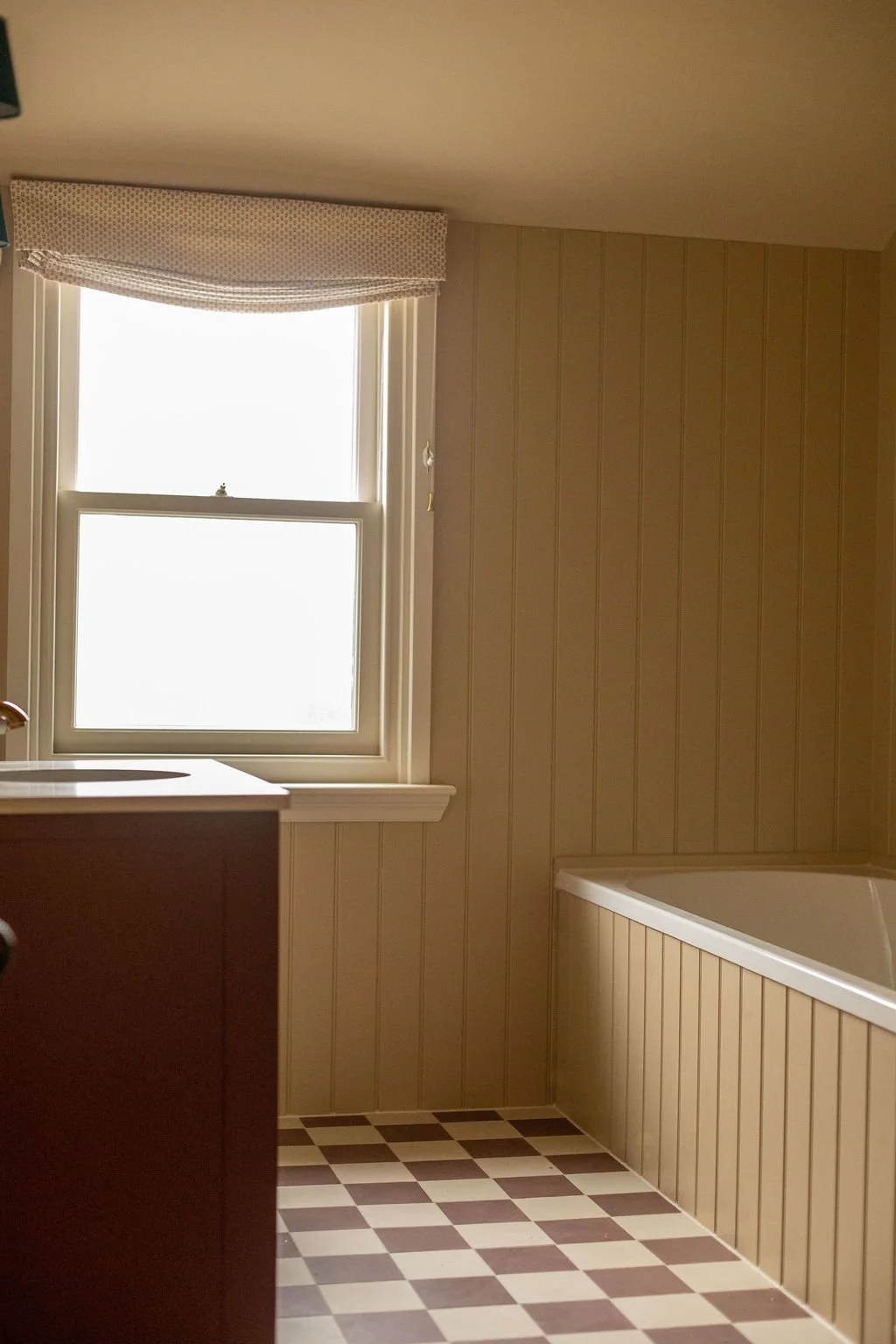 A bathroom with a window, checkered flooring, a bathtub, and cream paneled walls.