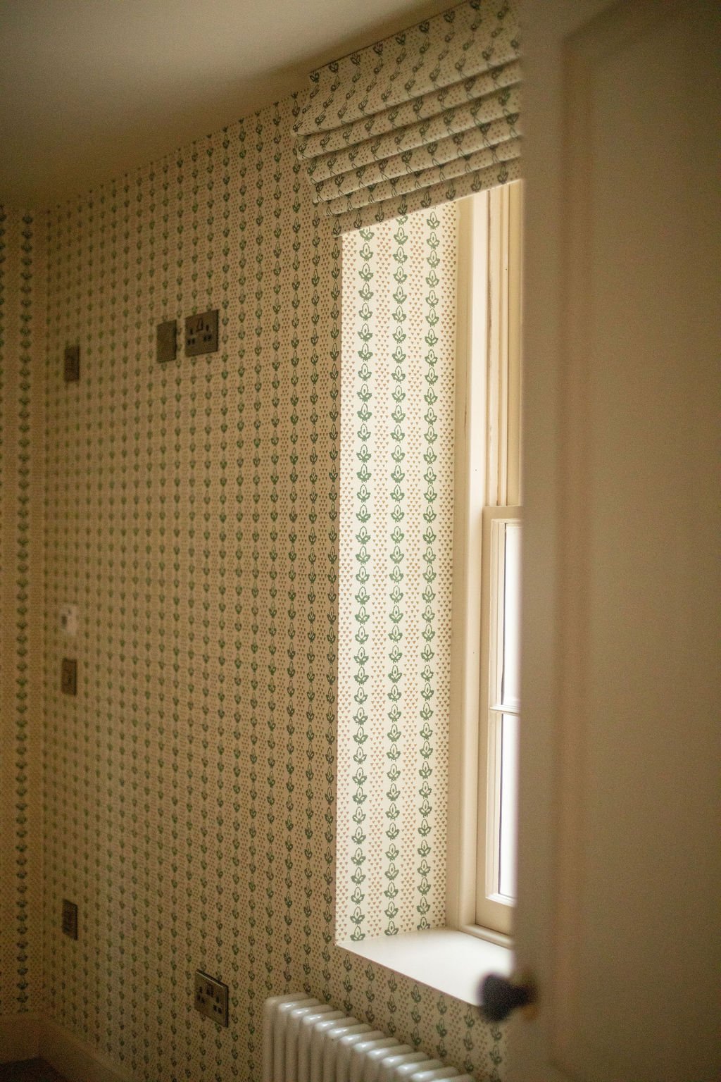 A corner of a room with patterned wallpaper, a window with a sheer curtain, a radiator, and switches on the wall.