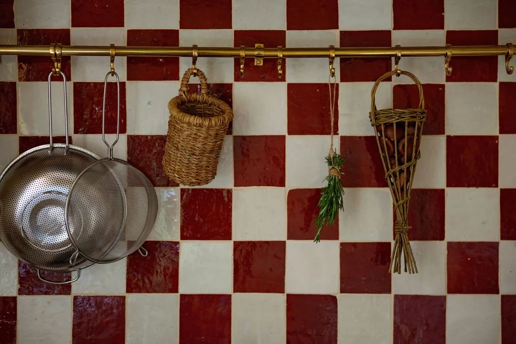 Kitchen wall with red and white checkered tiles, stainless steel strainers hanging on hooks, a woven basket, a bunch of herbs tied with string, and a wicker cone-shaped container.