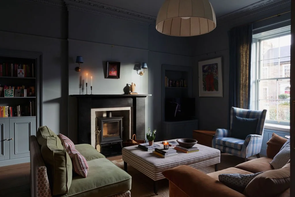 A cozy living room with a fireplace, blue bookshelf, armchair with plaid pattern, green and brown sofas, a large window with curtains, coffee table with books, and candles, in a traditional style.