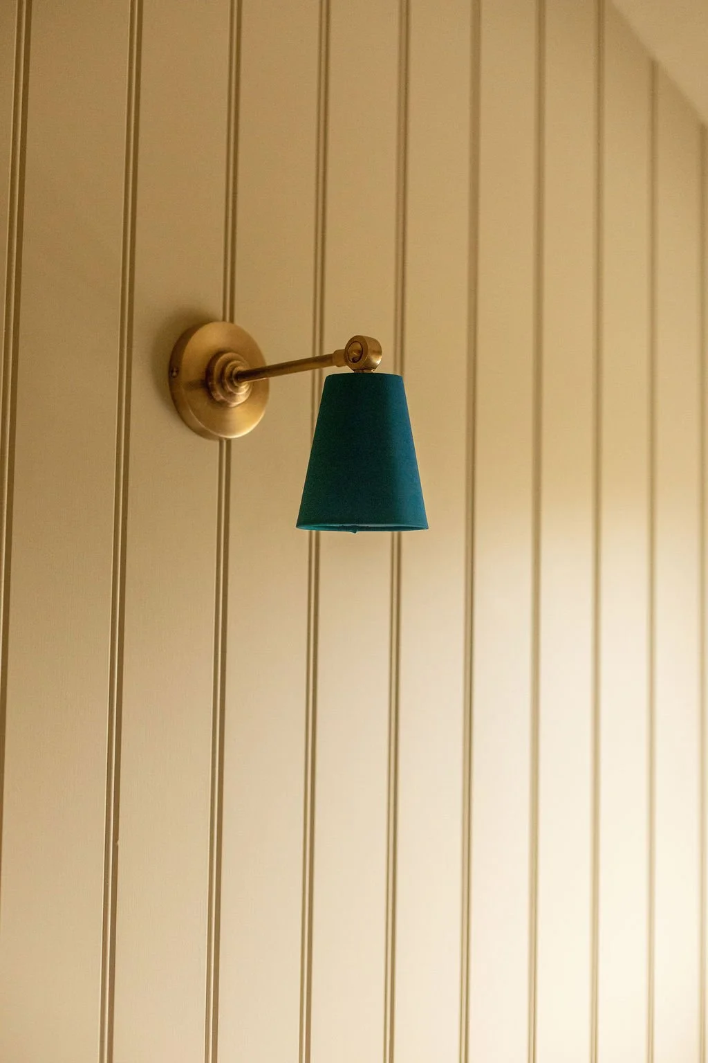 Wall-mounted lamp with a brass arm and a teal lampshade on a cream-colored vertical-paneled wall.