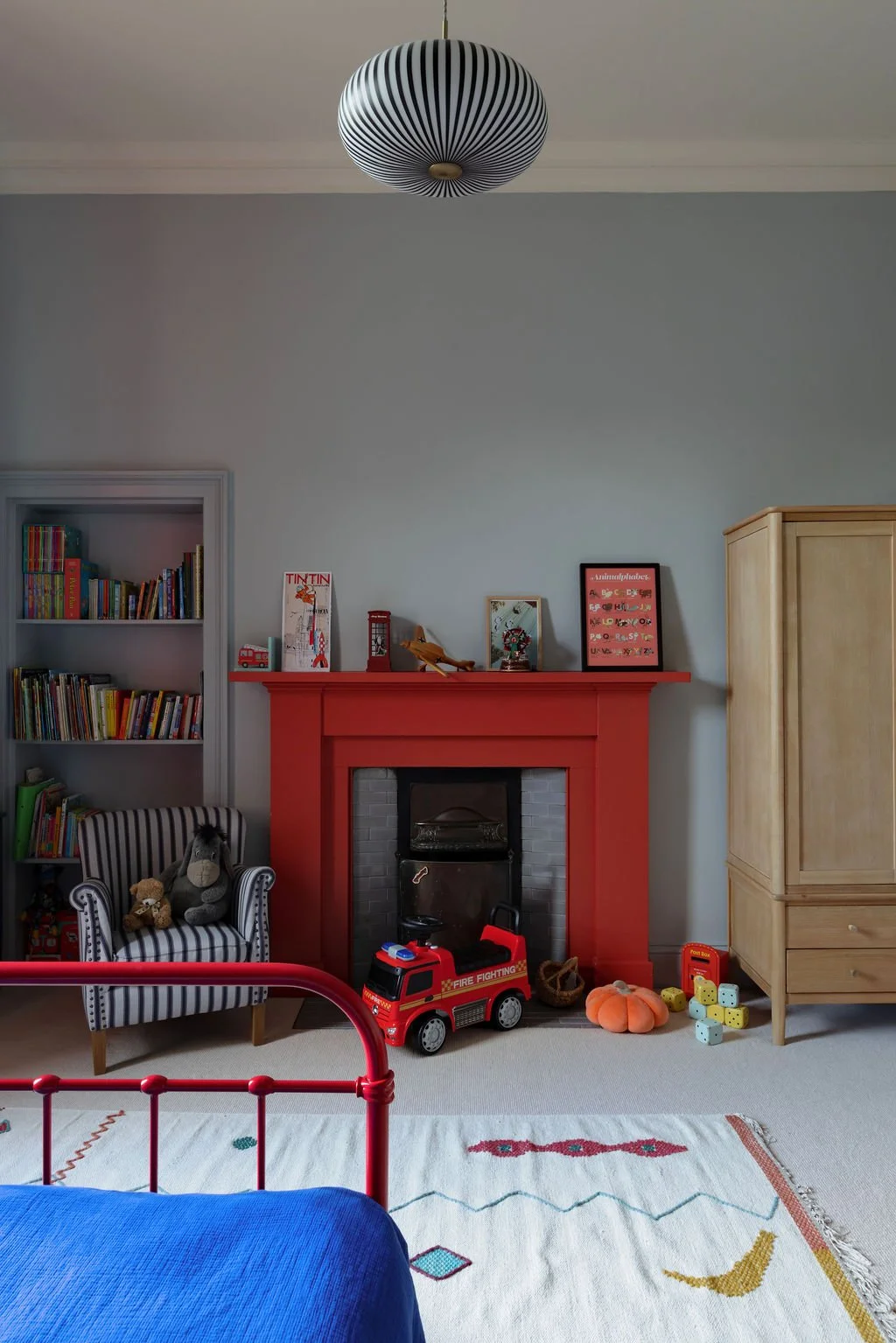 A cozy children's playroom featuring a red fireplace, a striped armchair with plush toys, a children's bed with a blue bedspread, a bookshelf filled with books, toys on the floor including a toy fire truck, dice, and a pumpkin-shaped pillow, and a ce
