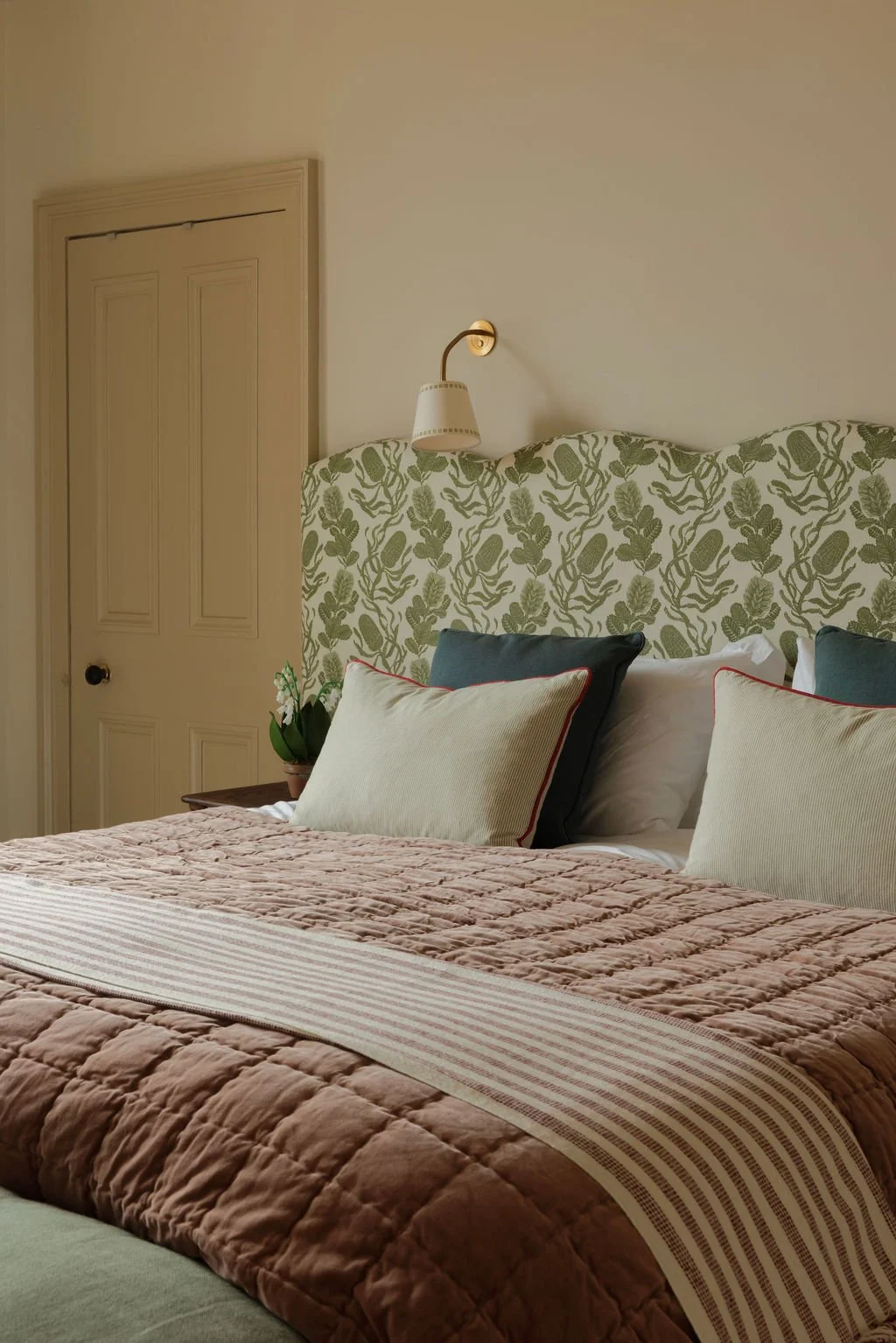 A cozy bedroom with a bed featuring a patterned green leafy headboard, pink quilted bedspread, and decorative pillows. A wall-mounted reading lamp is above the bed, and a small potted plant is on a bedside table. There's a closed door with a black do