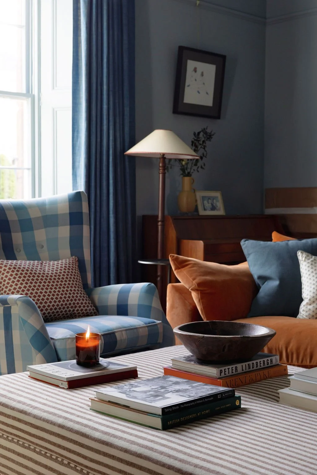Living room with blue checkered armchair, brown sofa with pillows, side table with lit candle, coffee table with books and large bowl, sideboard with lamp, framed photo, vase, window with blue curtains, framed artwork on blue wall.