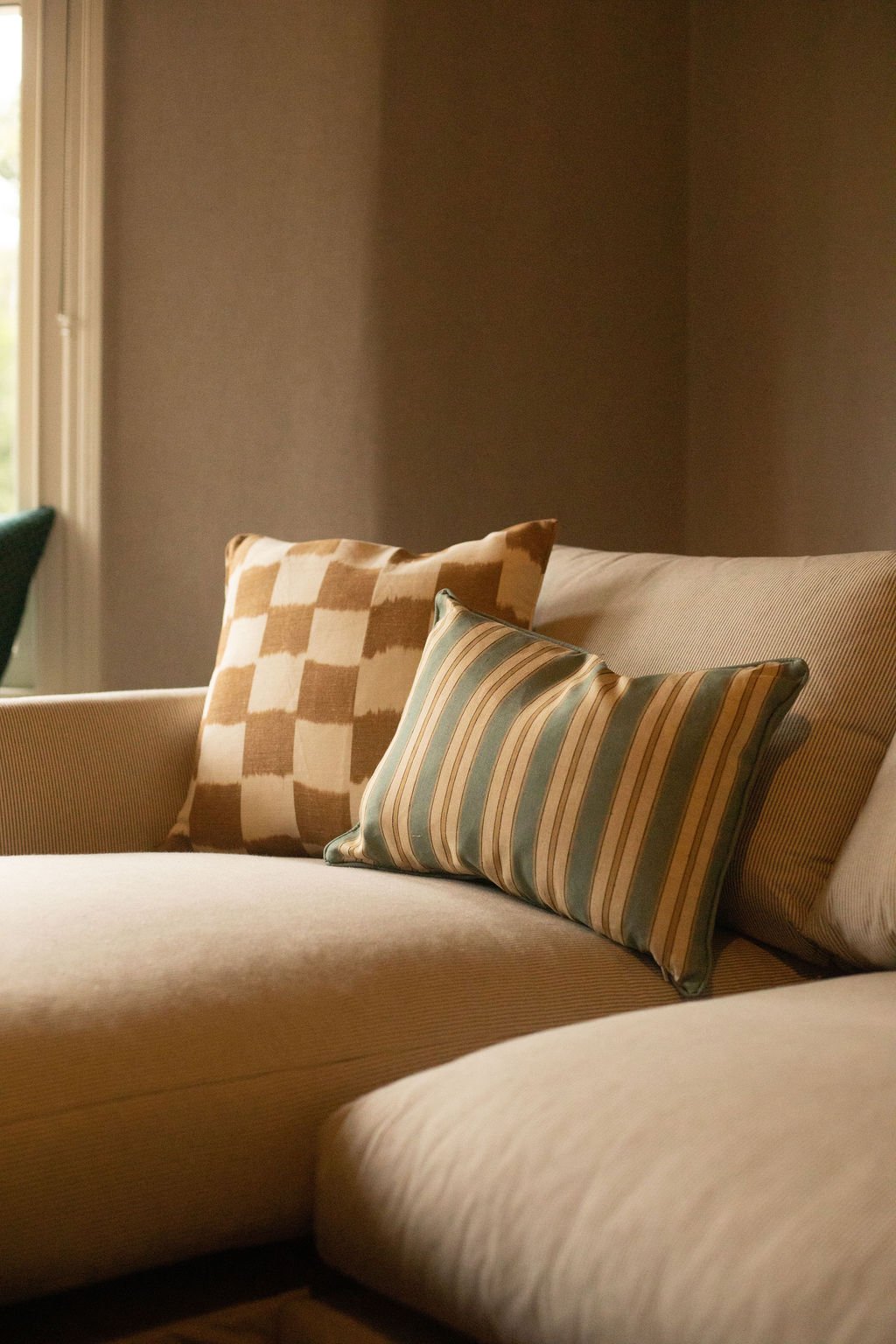 Close-up of a beige sofa with two decorative pillows, one with a checkered pattern and the other with striped pattern, near a window with natural light.