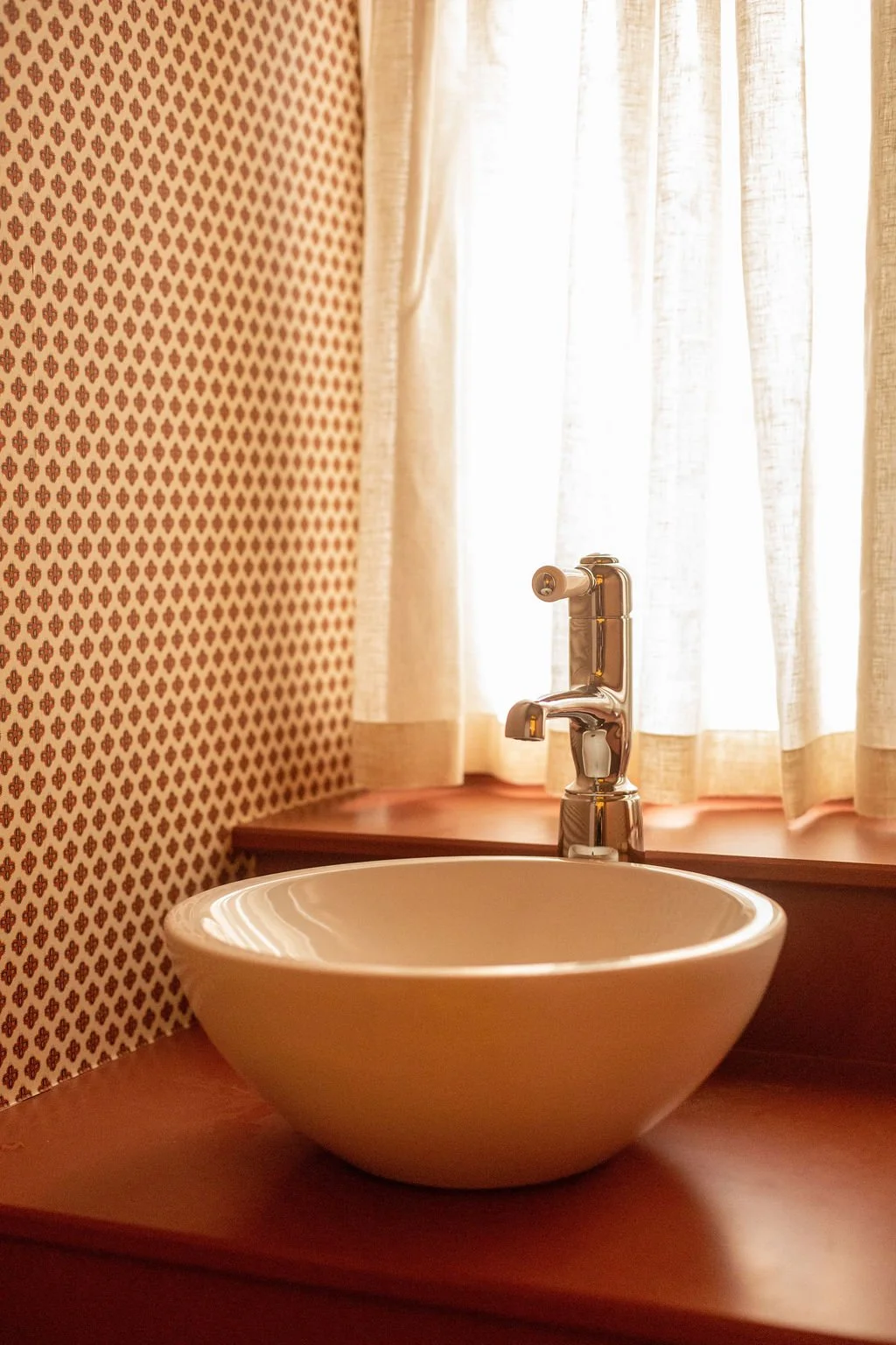 A white ceramic bowl sink with a chrome tap on a wooden counter, beside sheer curtains and patterned wallpaper, illuminated by soft light.