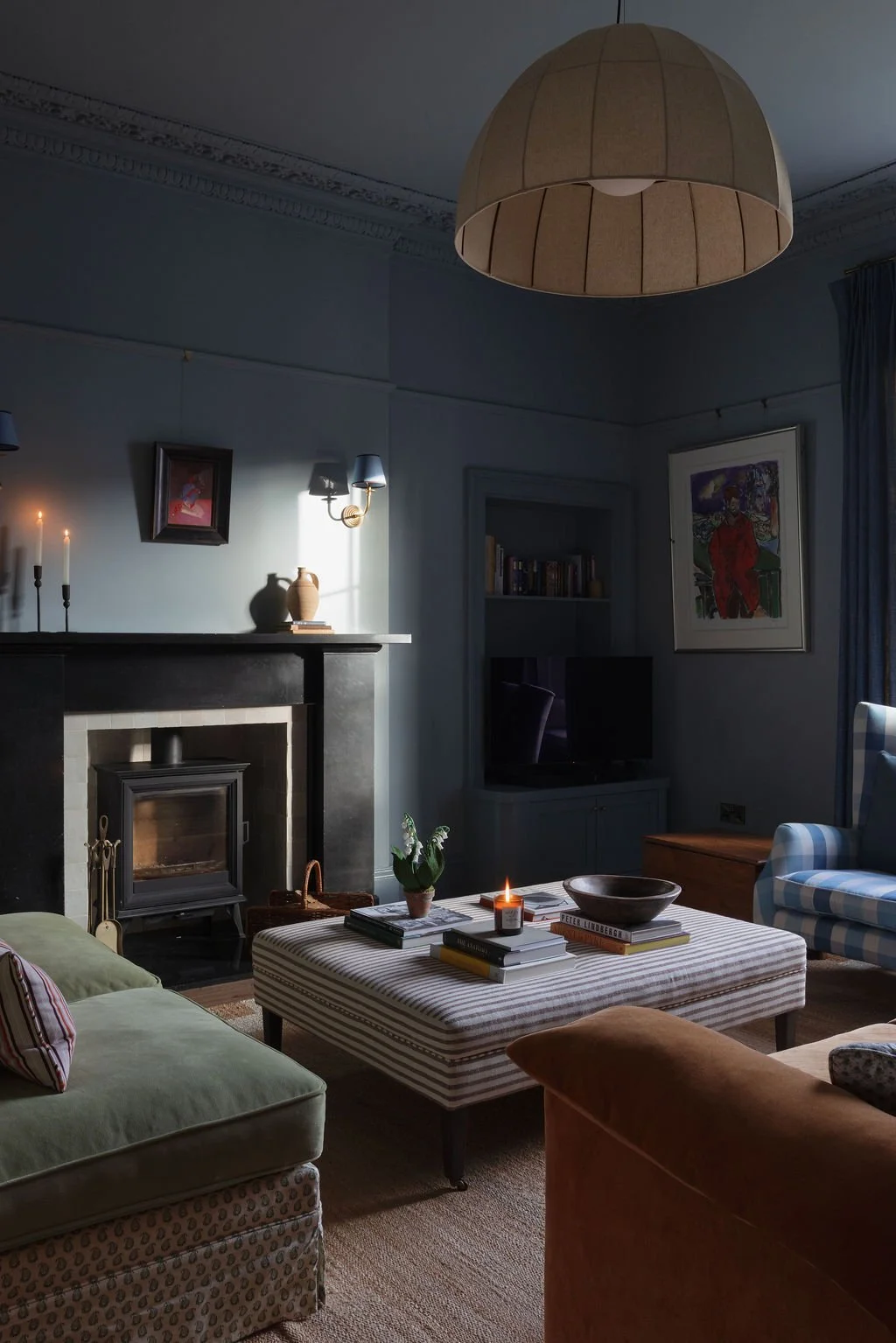 Cozy living room with a lit candle on a striped ottoman, a fireplace, a green sofa with a pillow, a blue and white checkered armchair, and a large framed painting on the wall.