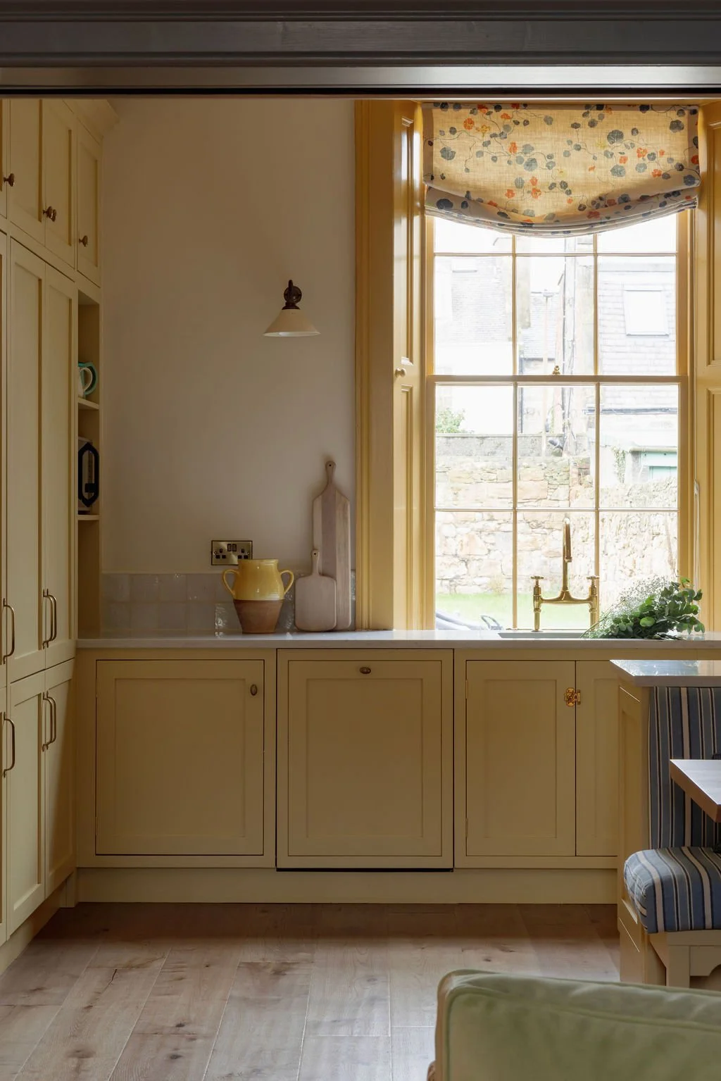 A cozy kitchen corner with a large window, yellow cabinetry, a white marble countertop, and decorative items including a yellow pitcher, a pink chopping board, and a plant near the sink.