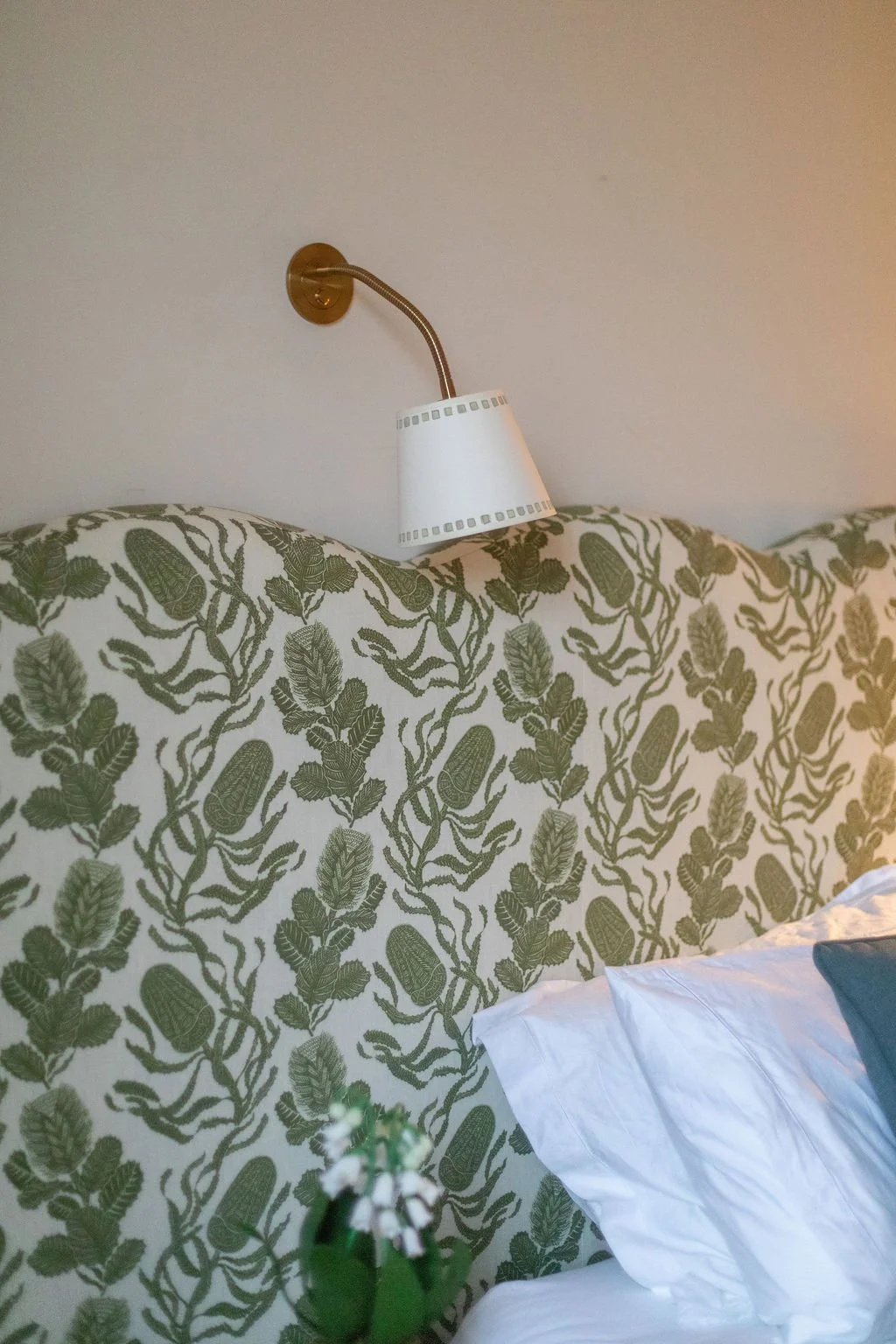 A bed with a patterned headboard featuring green leaves and plants, a white pillow, a small green plant in the foreground, and a brass wall-mounted reading light with a white lampshade.