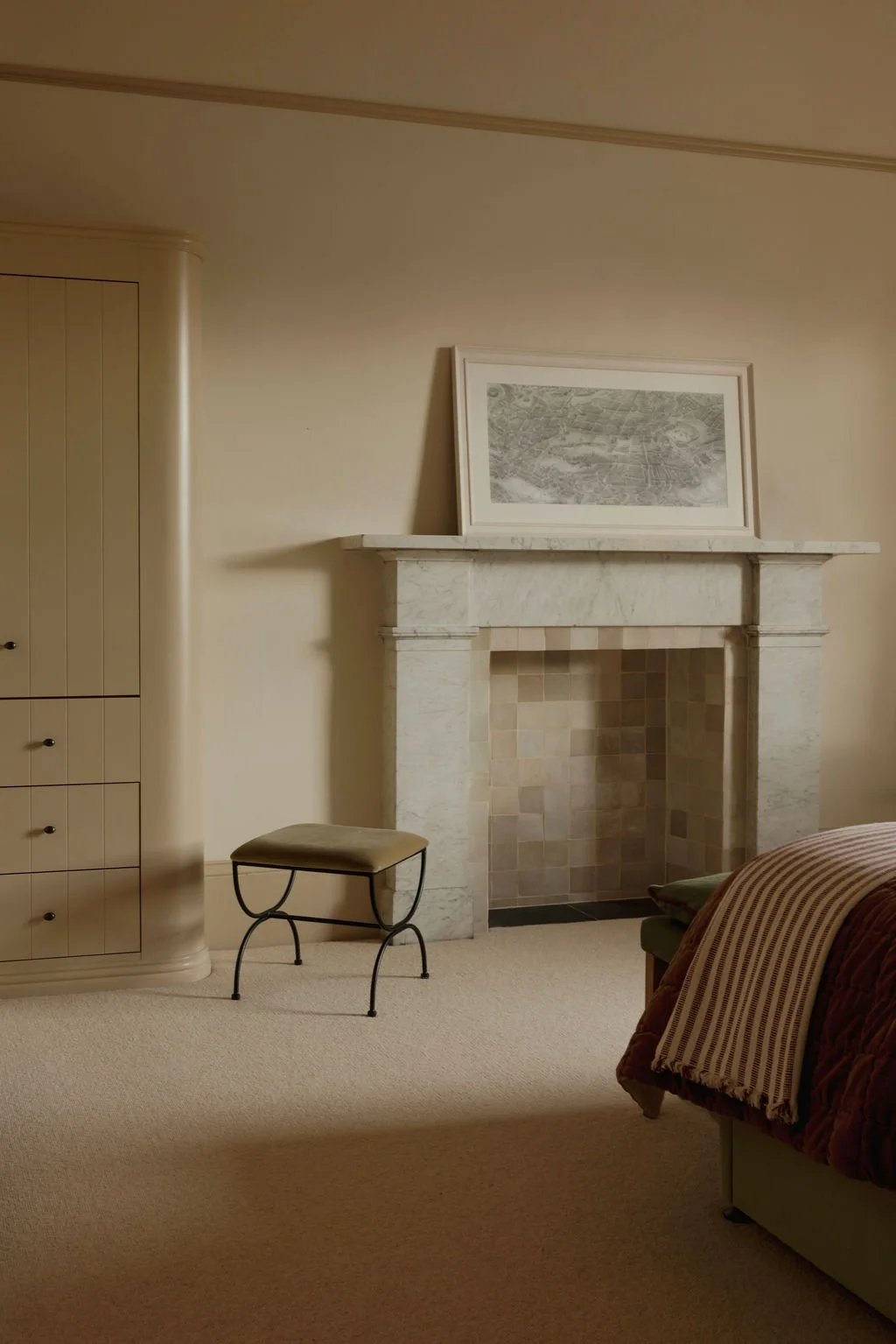 Living room with a white marble fireplace, framed map on mantel, cream walls, beige carpet, nearby chair, part of bed with striped blanket.