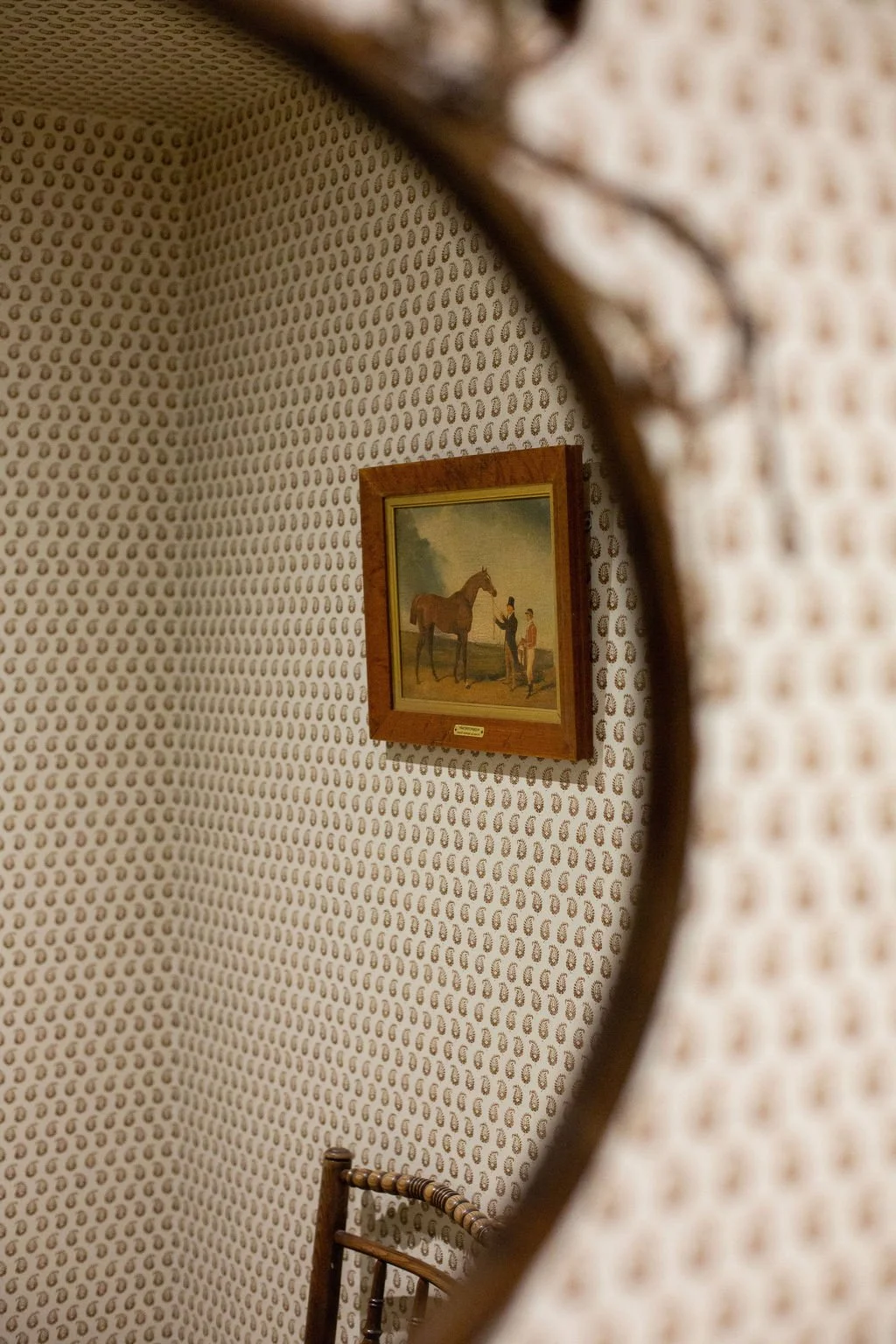 Reflection of a picture of a horse and two people hanging on an ornate wallpapered wall, seen through a mirror.