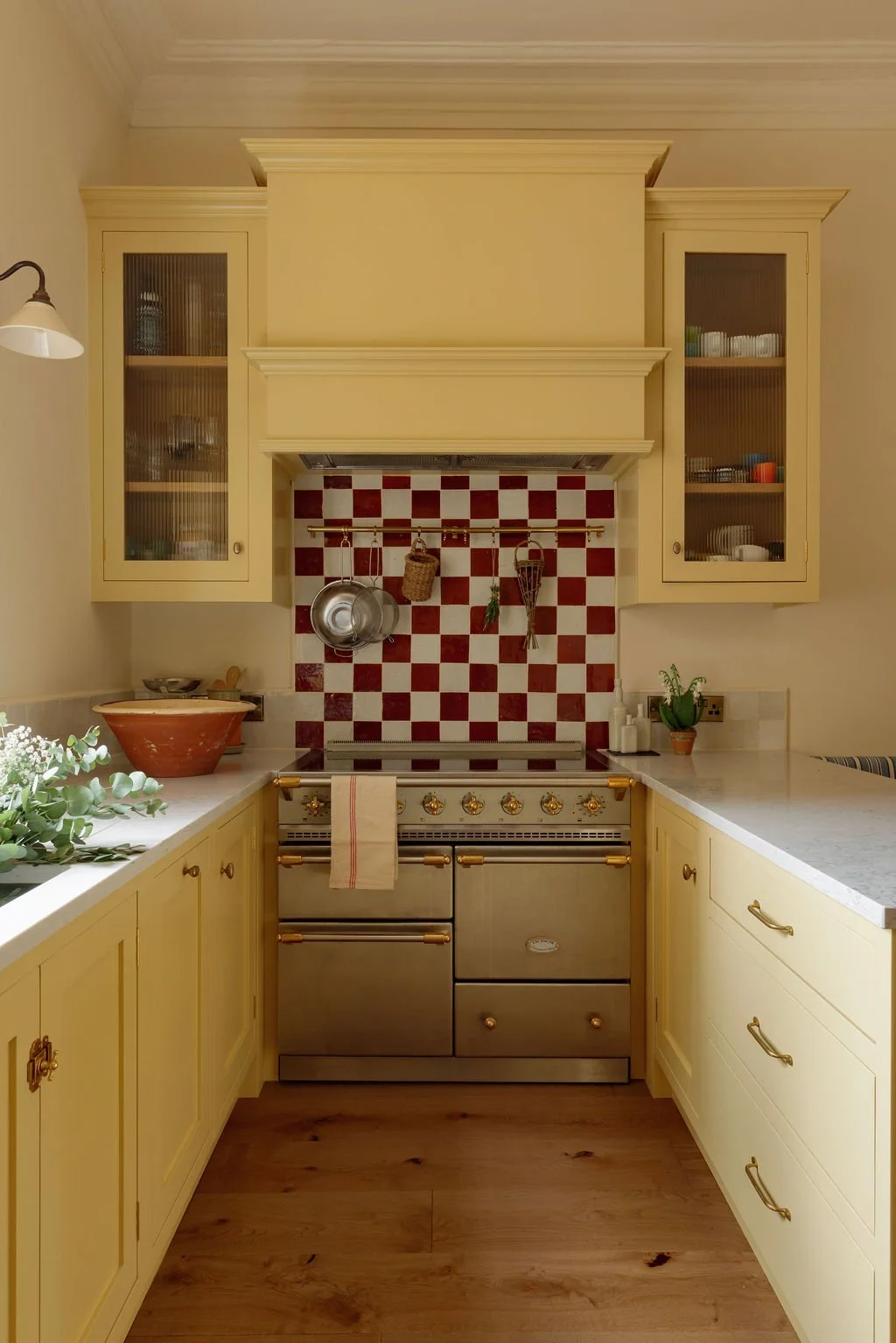 A cozy kitchen with yellow cabinets, a red and white checkered tile backsplash, a vintage oven, and decorative items including potted plants and hanging kitchen utensils.