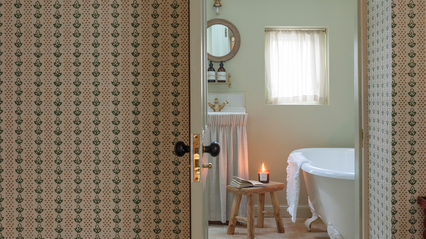 View through a door into a vintage bathroom with a clawfoot tub, a small wooden stool with an open book and a lit candle, a sink with a gold faucet, framed mirror, vanity with toiletries, and a window with sheer curtains.