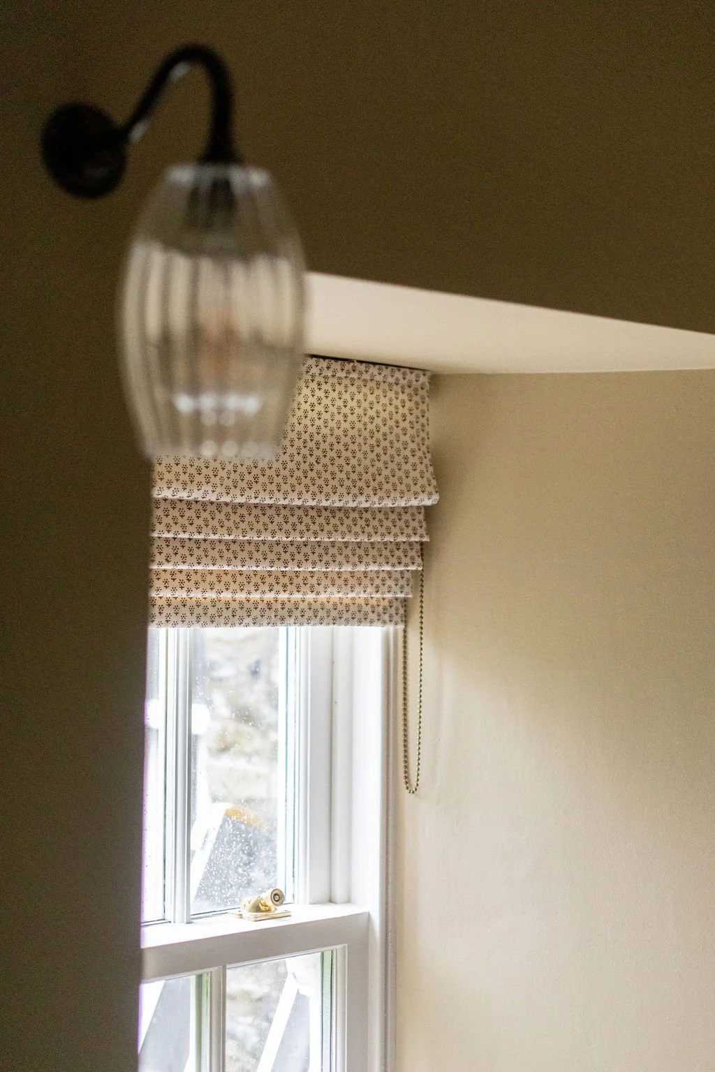 Close-up view of a decorative window shade with a small bead chain, partially covering a window with a natural landscape outside.