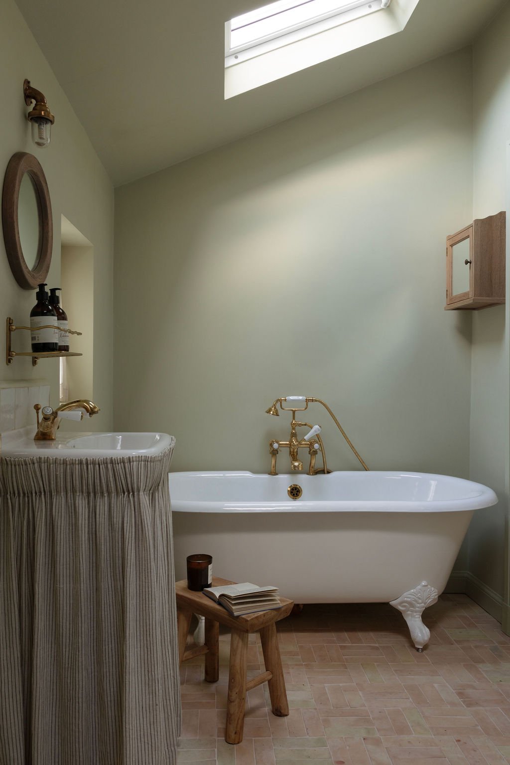 Bathroom with a white clawfoot bathtub, a small wooden stool with an open book and a candle, a pedestal sink with a striped curtain, and a skylight overhead.