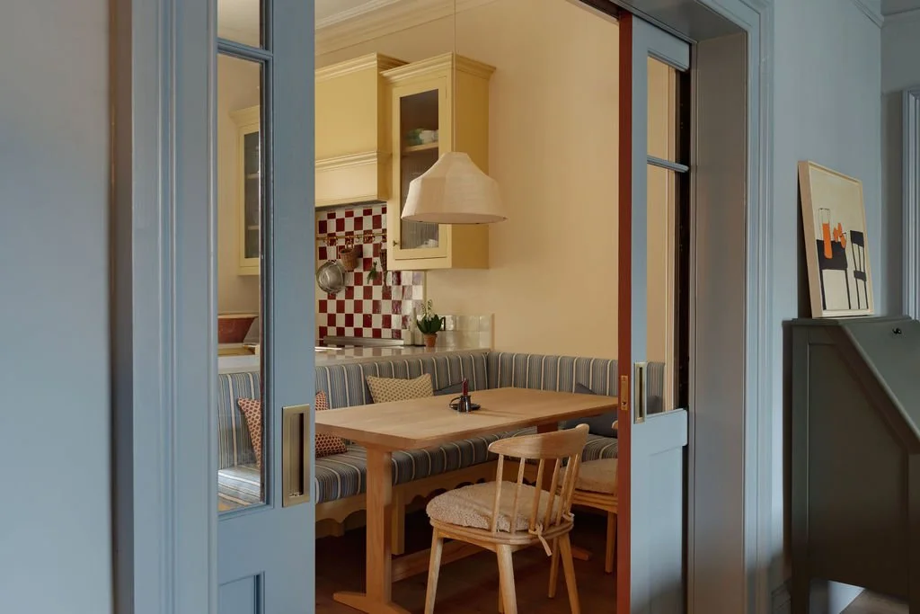 View of a cozy dining nook with a wooden table, striped cushioned bench, and wooden chairs, seen through a blue sliding door in a kitchen featuring yellow cabinetry and red checkered backsplash.