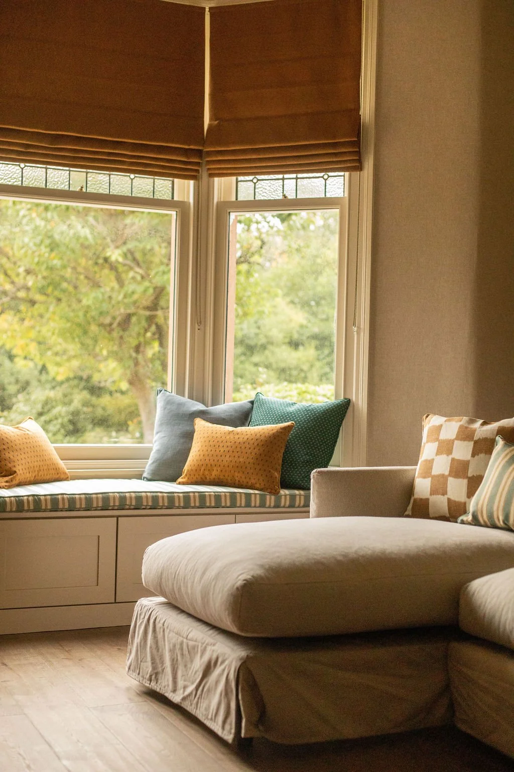 Sunlit living room with window seat and beige sofa, decorated with colorful pillows, overlooking trees outside.