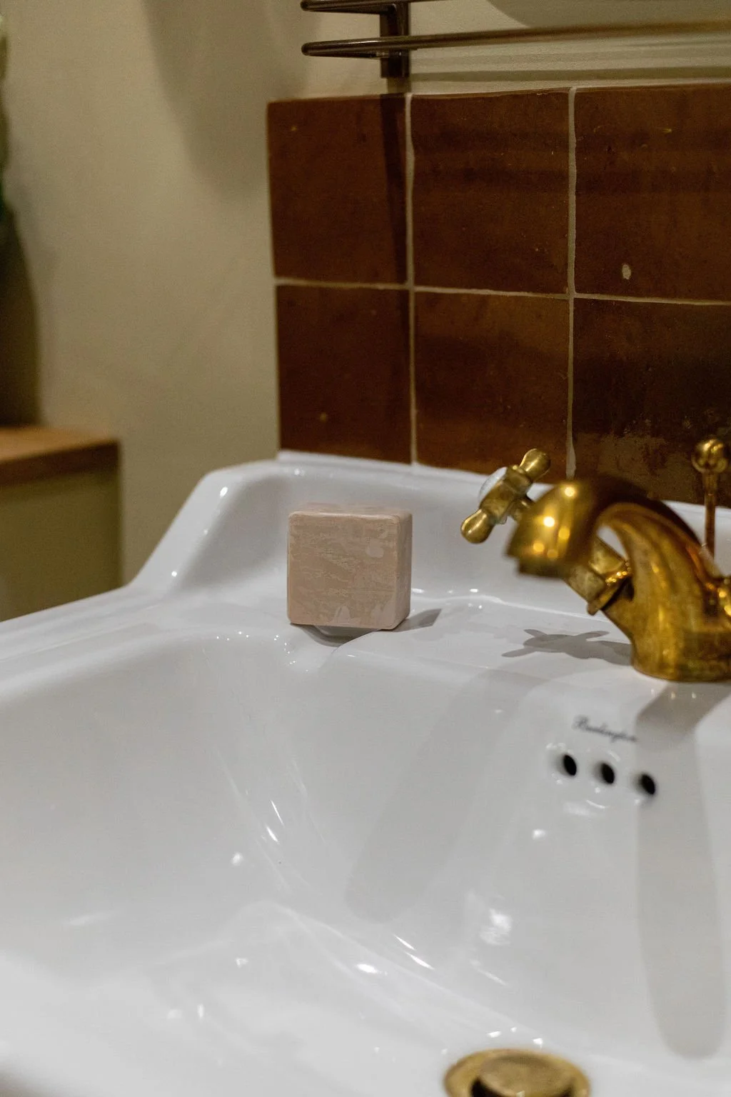 A white bathroom sink with a gold faucet, brown tiled wall in the background, and a small beige soap bar on the sink. A soap dish is partially visible on the left.