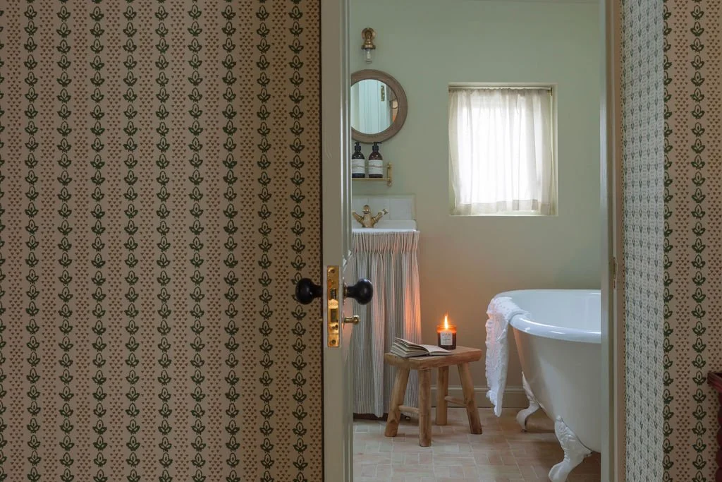 View into a bathroom through a partially open door. The bathroom contains a clawfoot tub, a small wooden stool with a lit candle and an open book, a sink with a striped skirt, a mirror, and a window with sheer curtains.