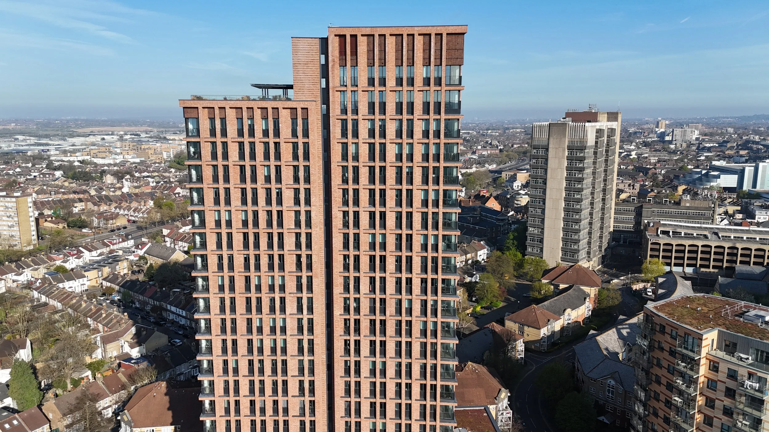 A tall modern residential building with a brick facade, featuring balconies and narrow windows, surrounded by a mix of older houses and other high-rise buildings in an urban area under a clear blue sky.