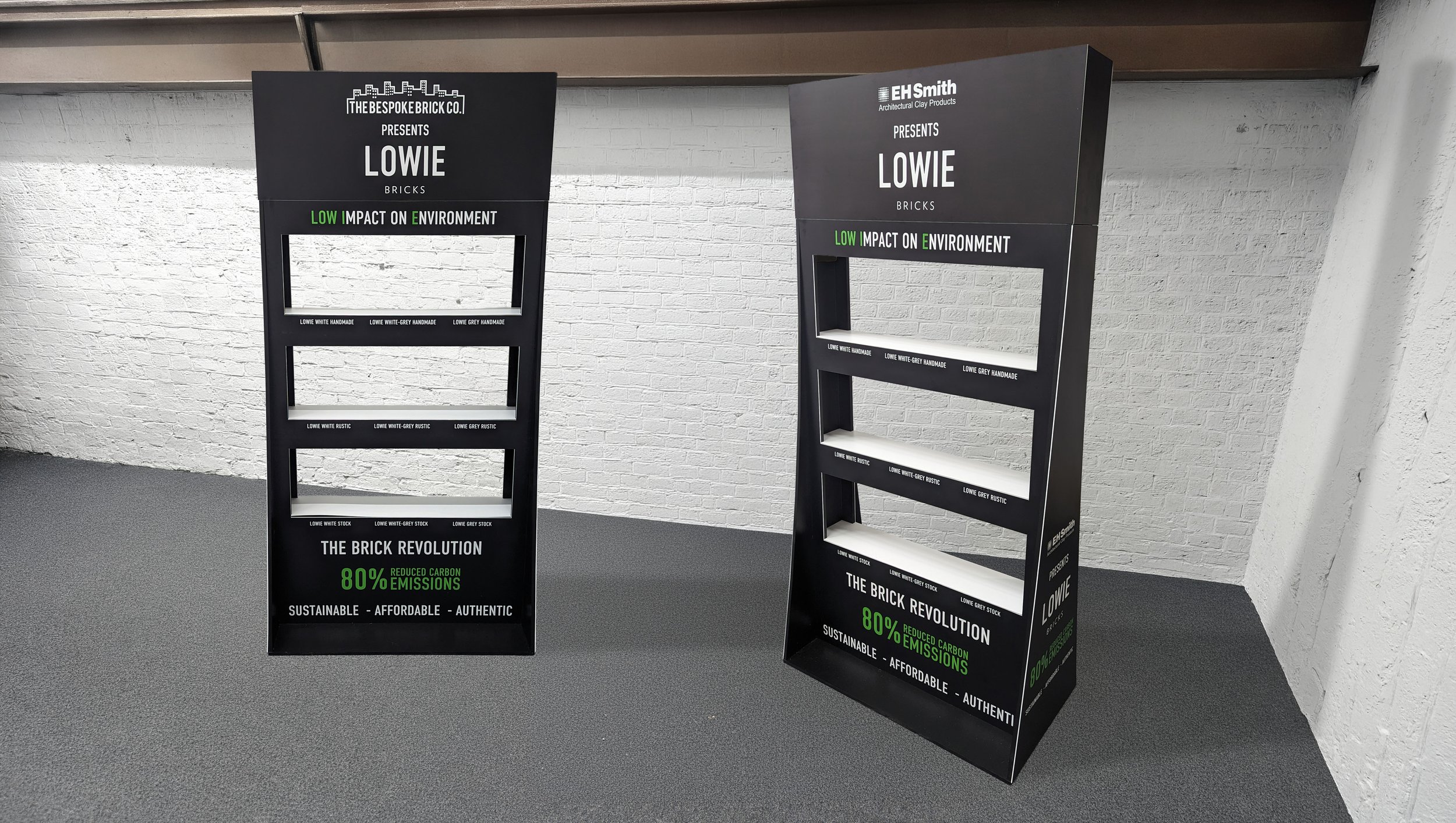 Two black display stands promoting eco-friendly bricks with the branding 'Lowie Bricks' and messages about low impact on the environment, reduced carbon emissions, and sustainability, set against a white brick wall in an indoor space.