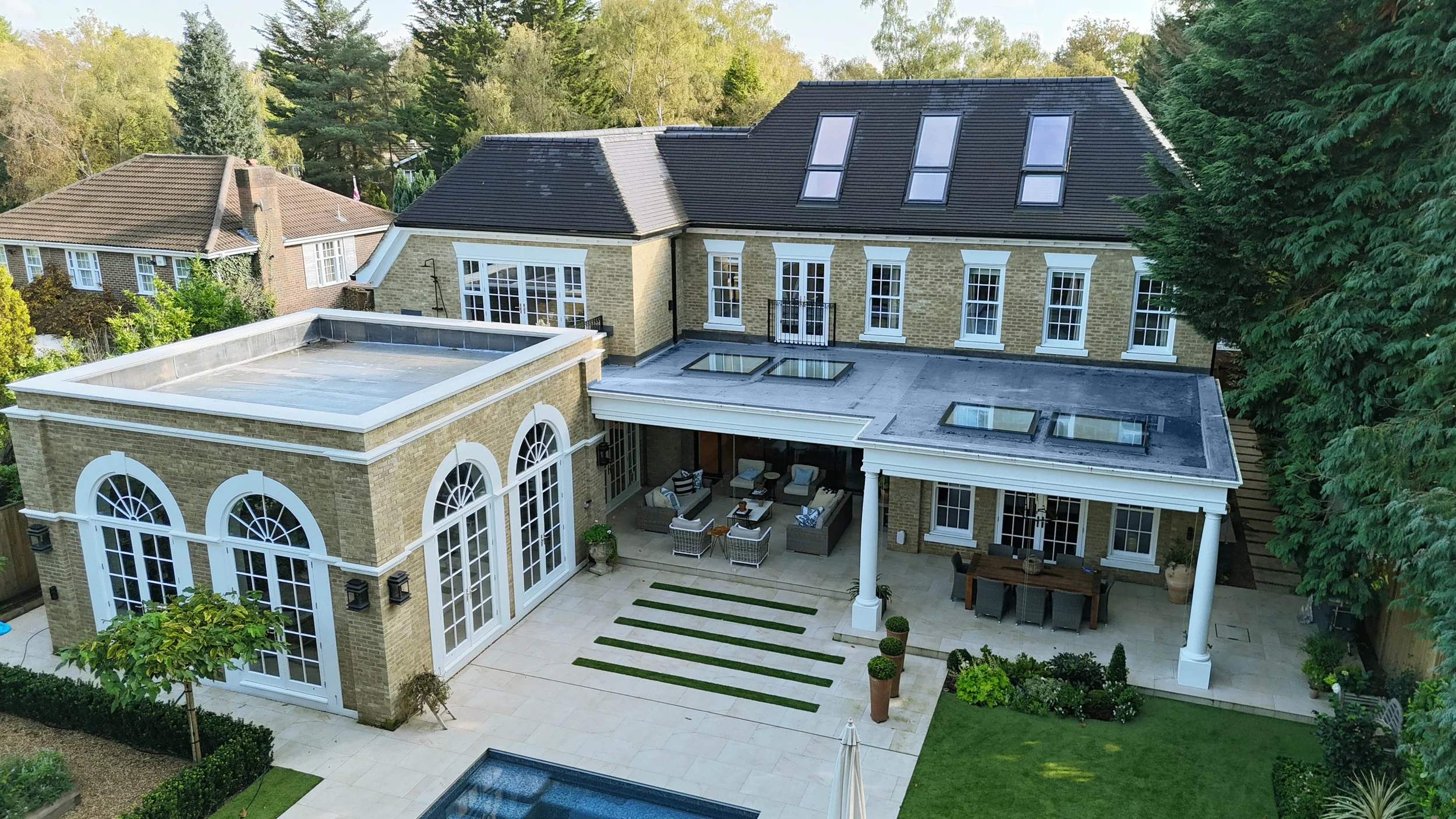 Aerial view of a large house with a landscaped backyard featuring a patio with outdoor furniture, a small pool, and a lush green lawn, surrounded by trees.