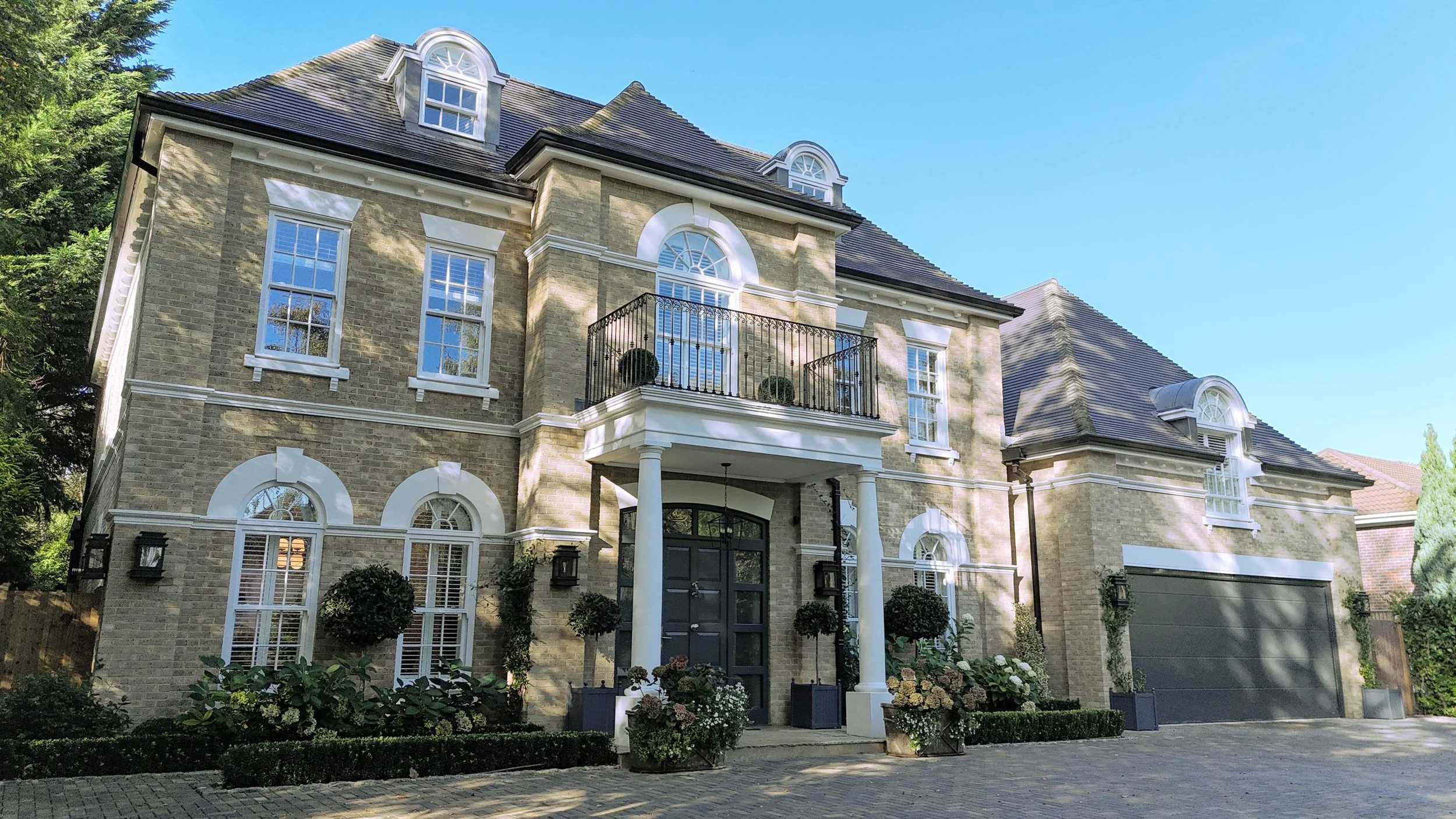 Large, elegant house with brick exterior, multiple tall windows, a small balcony, and a front entrance with columns, surrounded by landscaped garden with shrubs and trees.