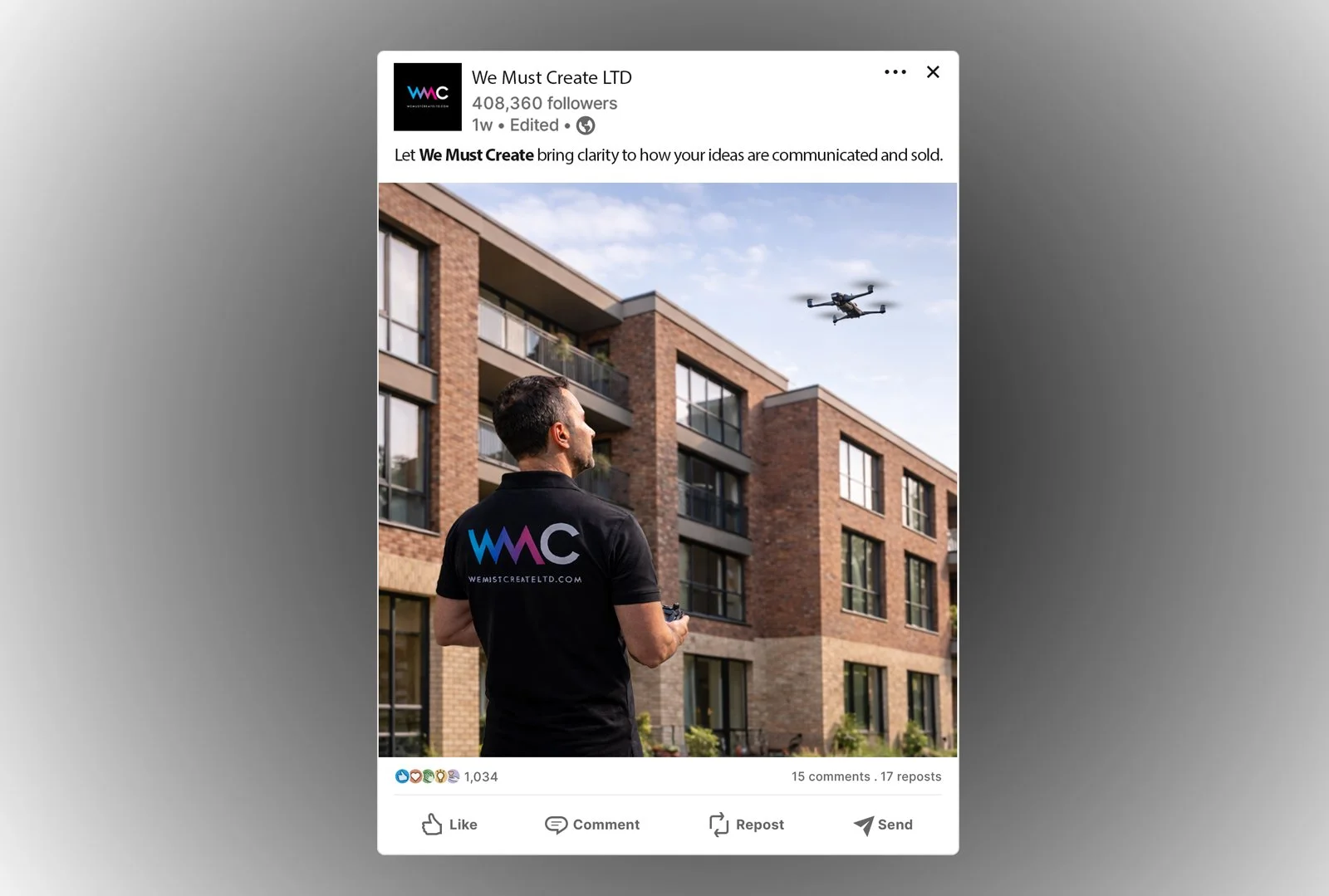 A man wearing a black polo shirt with a colorful company logo on the back is controlling a drone flying in the sky in front of a modern brick apartment building.