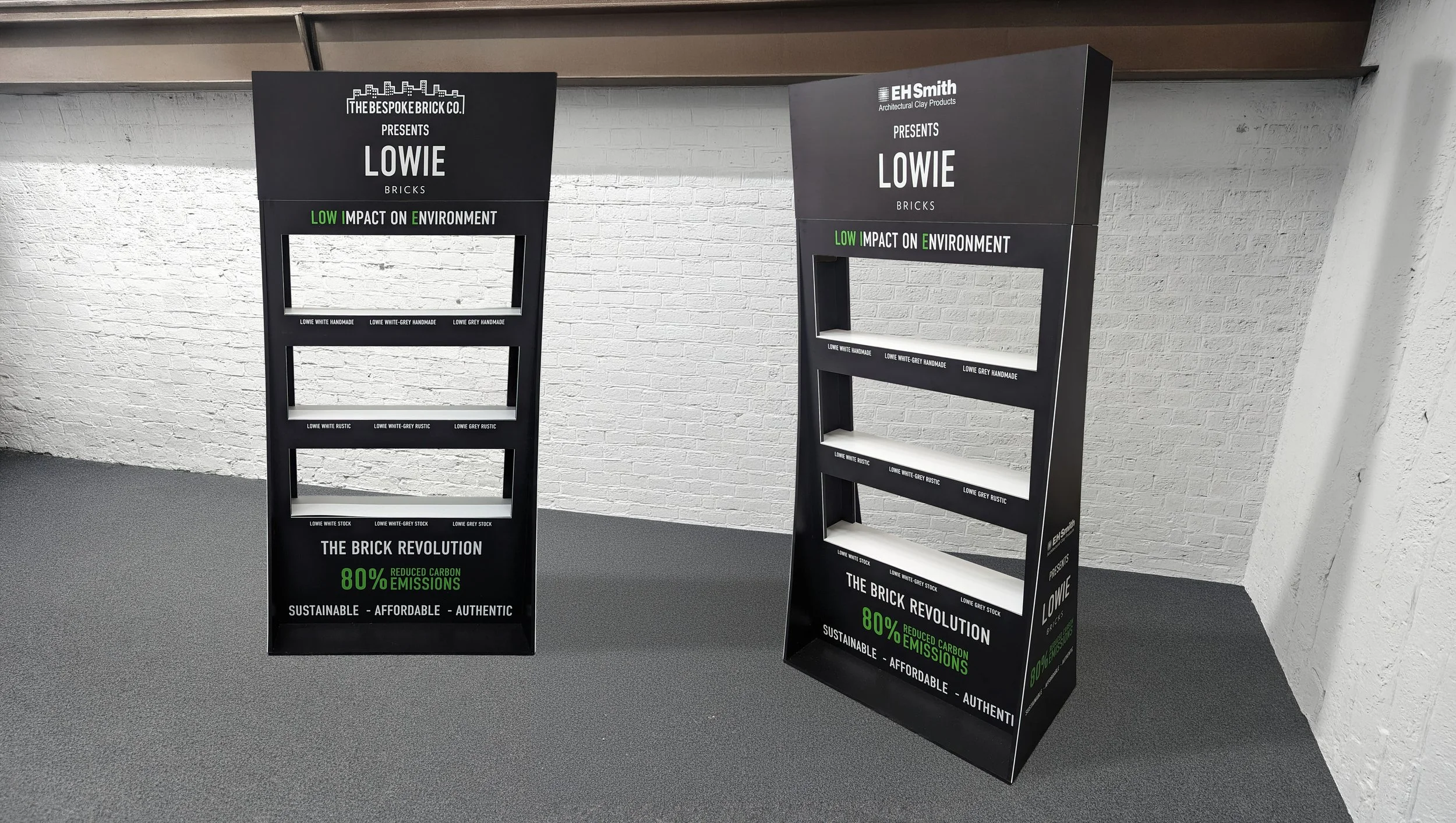 Two display stands promoting eco-friendly bricks with slogans about low impact on the environment, reduced carbon emissions, and sustainability, placed on a gray carpeted floor against a white brick wall.