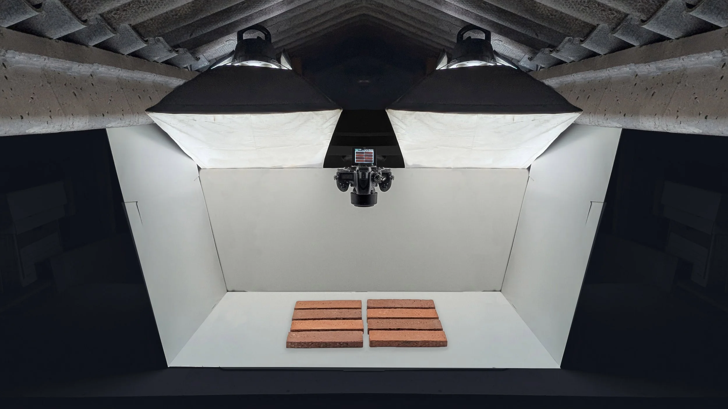 A studio photography setup with a camera on a tripod facing a white backdrop. There are three bricks placed on the floor in front of the backdrop, and two large studio lights with softboxes illuminate the scene. The setting appears to be in a dark room with black walls and ceiling.