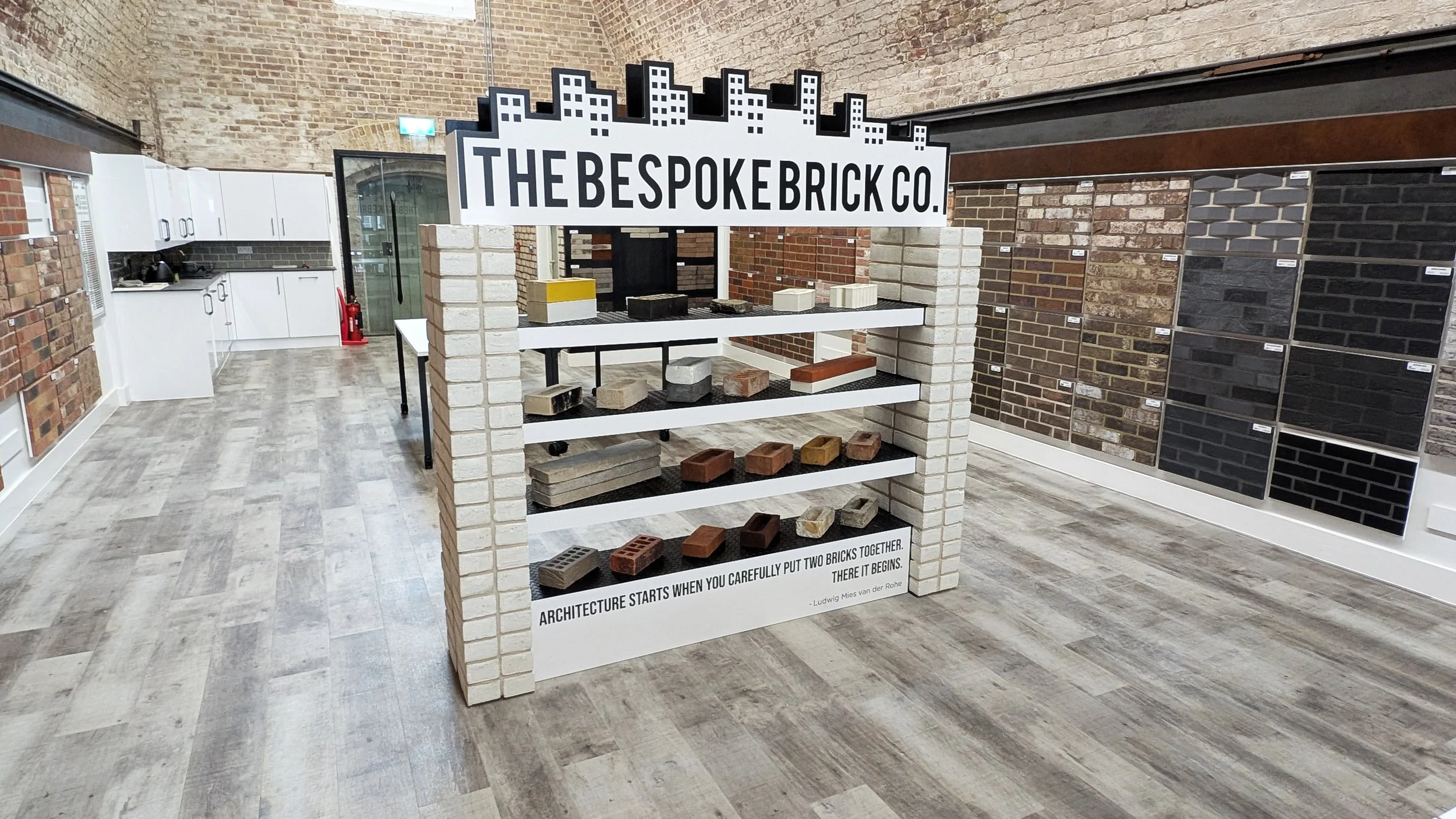 Display of various bricks and bricks samples on shelves in a showroom with a sign that reads 'The Bespoke Brick Co.' and a quote, 'Architecture starts when you carefully put two bricks together.'