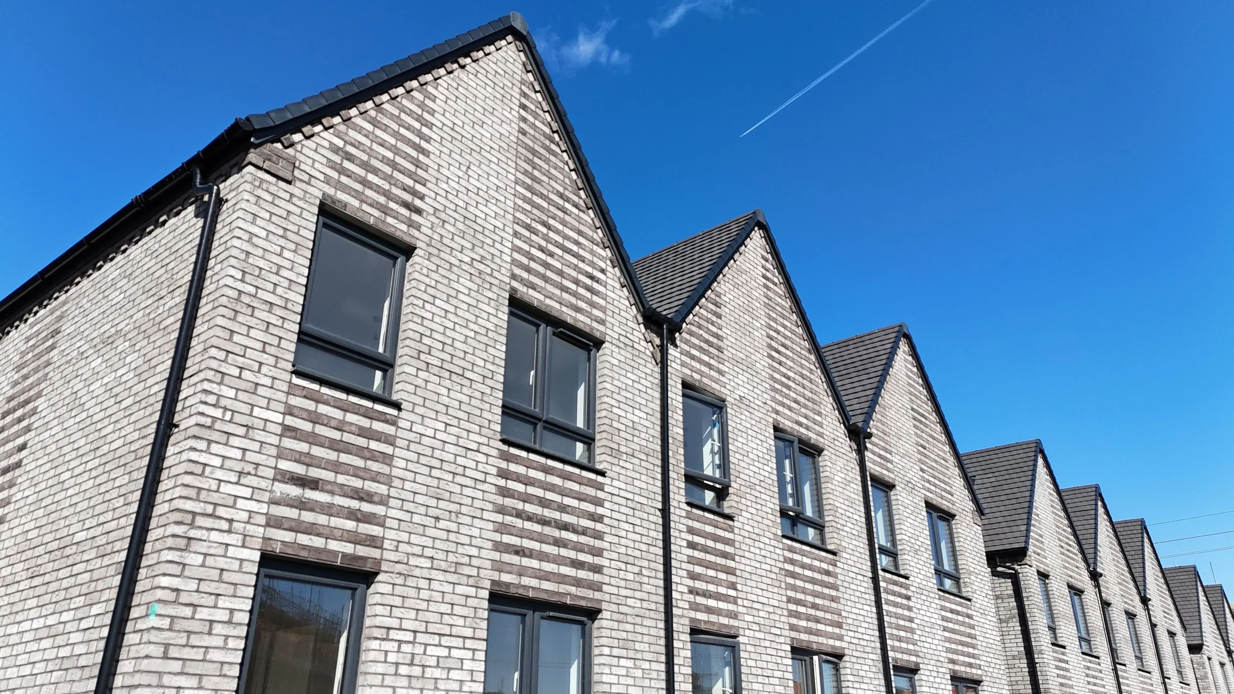 A row of modern brick townhouses with dark roof shingles, black window frames, and black gutter pipes under a clear blue sky with a visible contrail.