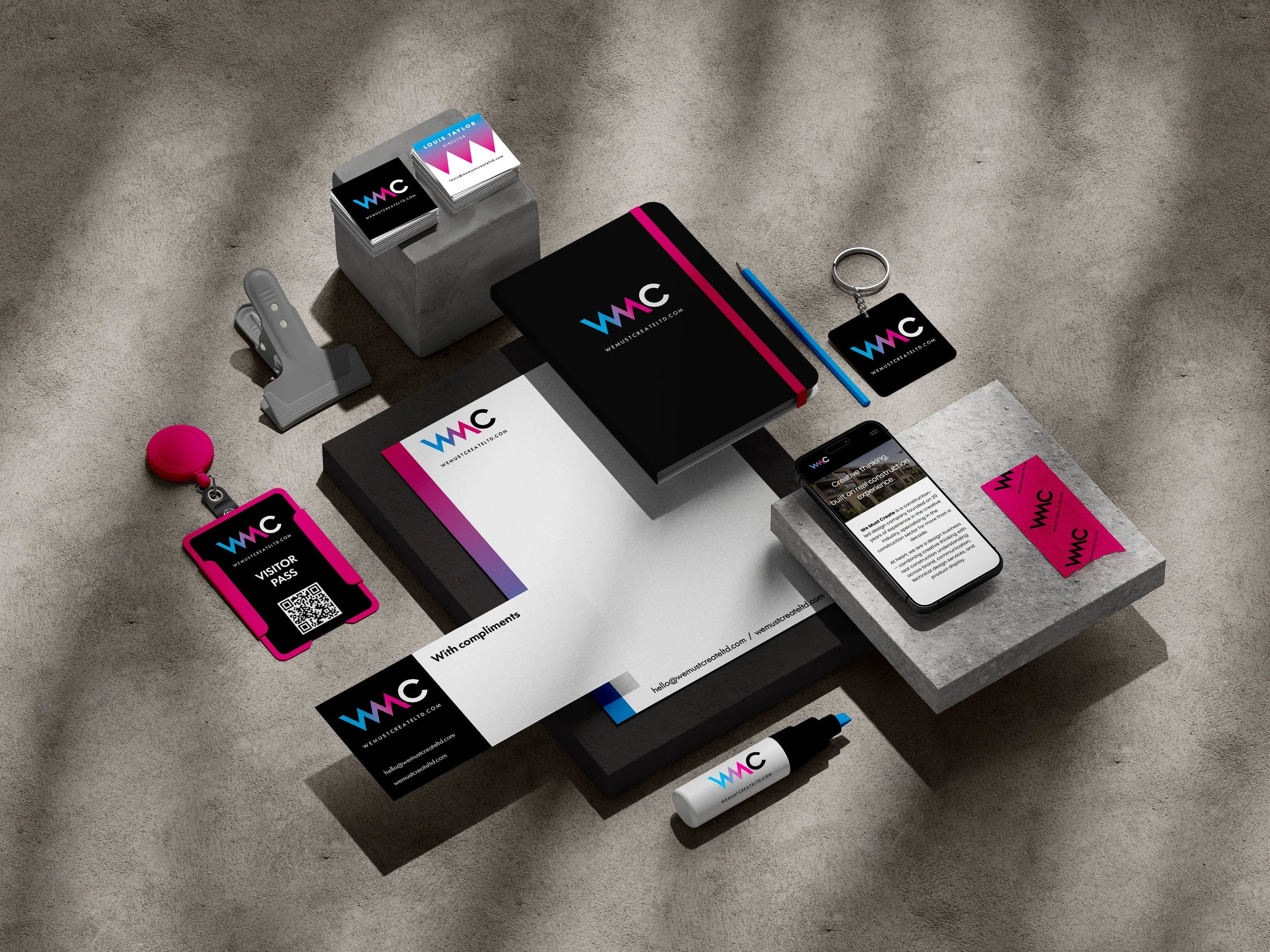 A variety of branded office supplies and promotional materials for WemusCreateLtd, including business cards, a notebook, a USB drive, a phone with a website, a name badge, a pen, and a marker, arranged on a textured surface.