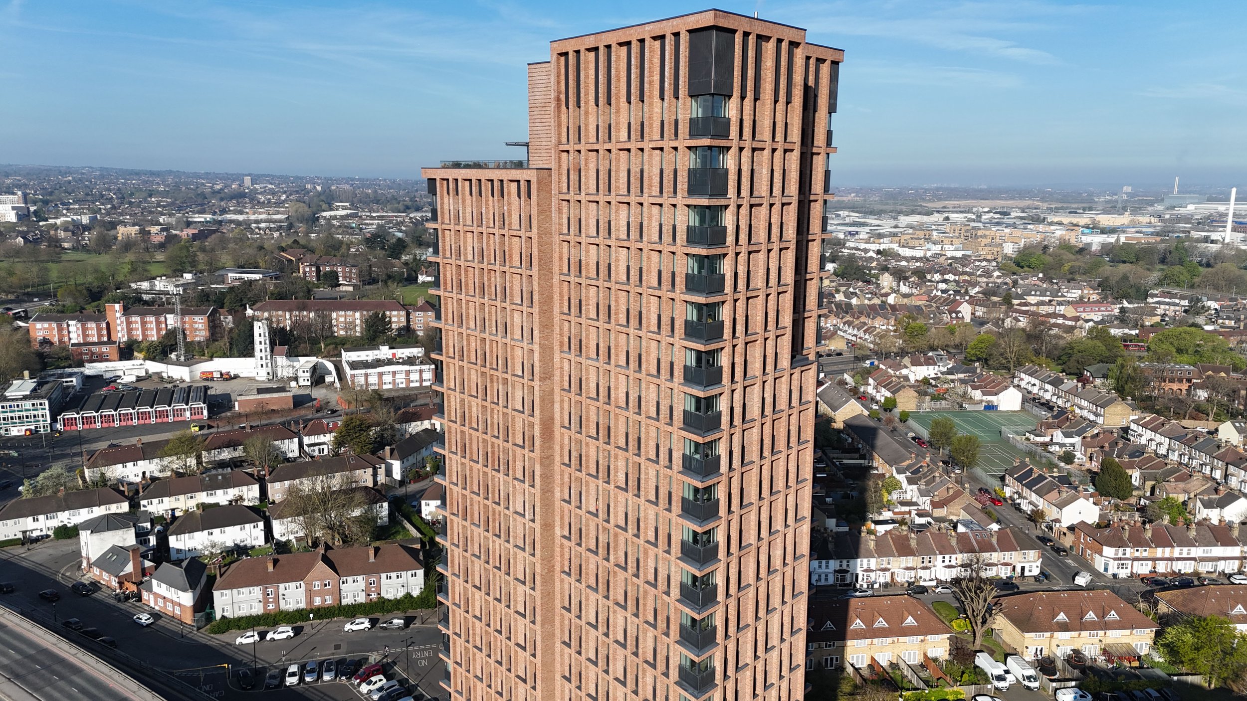 A tall, modern residential building with a brick exterior, featuring balconies with black railings on each floor, set against a backdrop of a cityscape with houses, commercial buildings, and distant greenery.