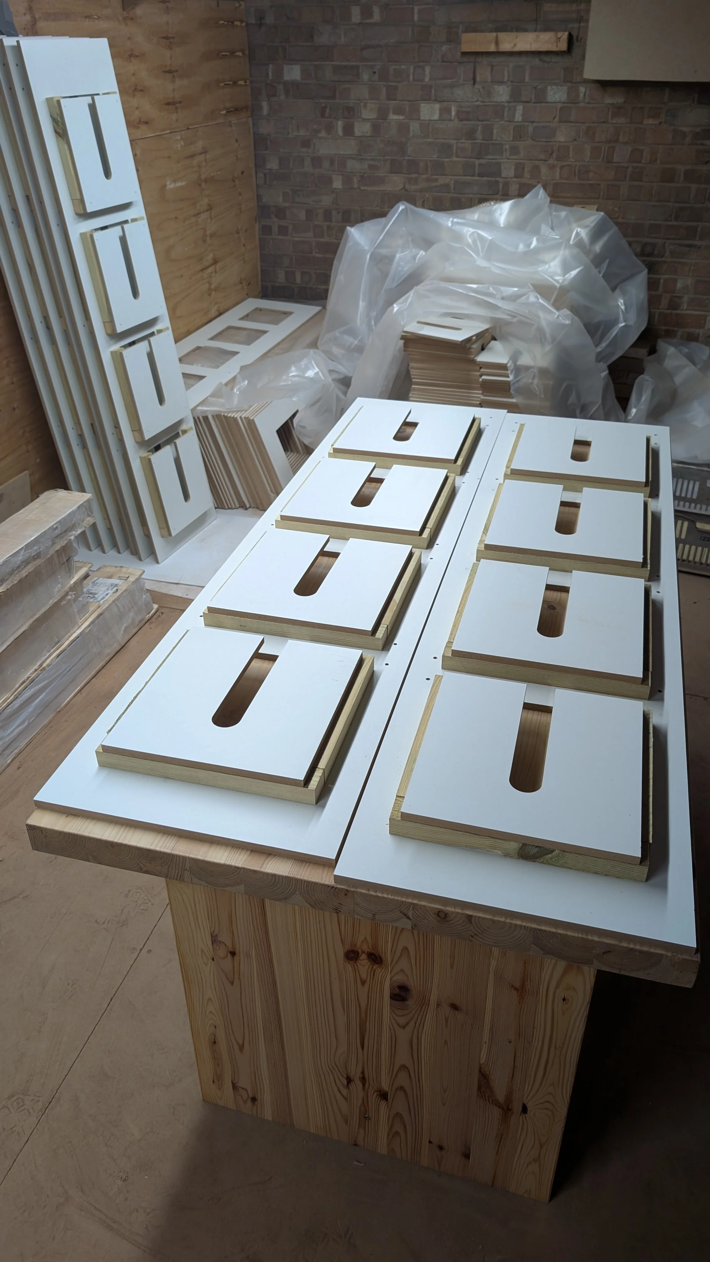 Multiple white cabinet door facades with cutouts, stacked and laid on a wooden workbench in a workshop setting with brick and wood walls.