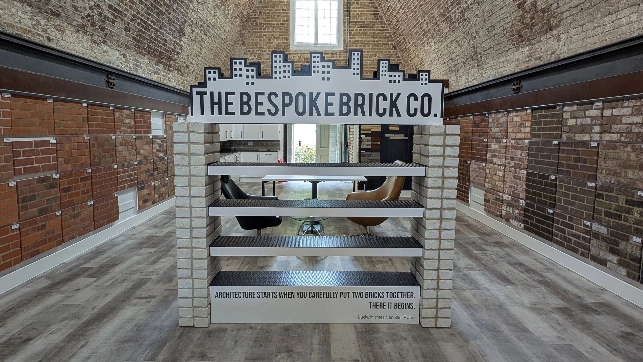 Interior of a brick showroom featuring display samples of various brick styles on the walls. A central brick structure with shelves displays more bricks, and a quote about architecture at the bottom.