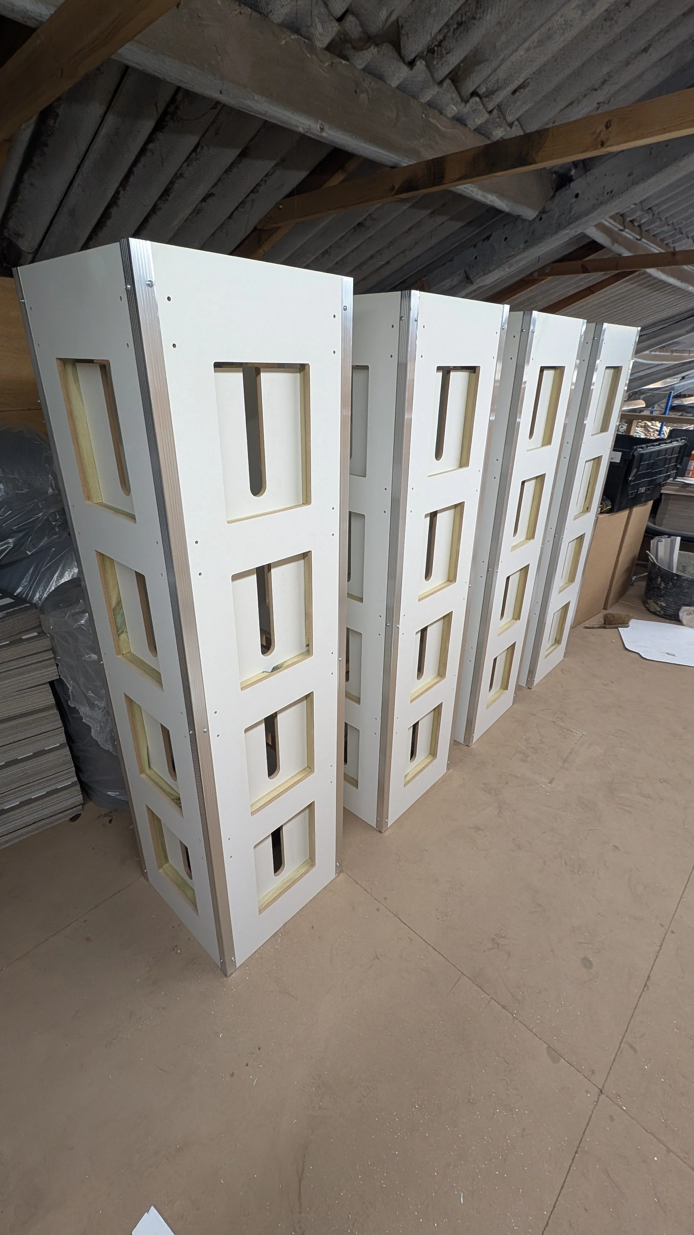 Several tall, white wooden frames with rectangular cutouts, standing upright in a workshop or storage area with unfinished walls and a concrete floor.
